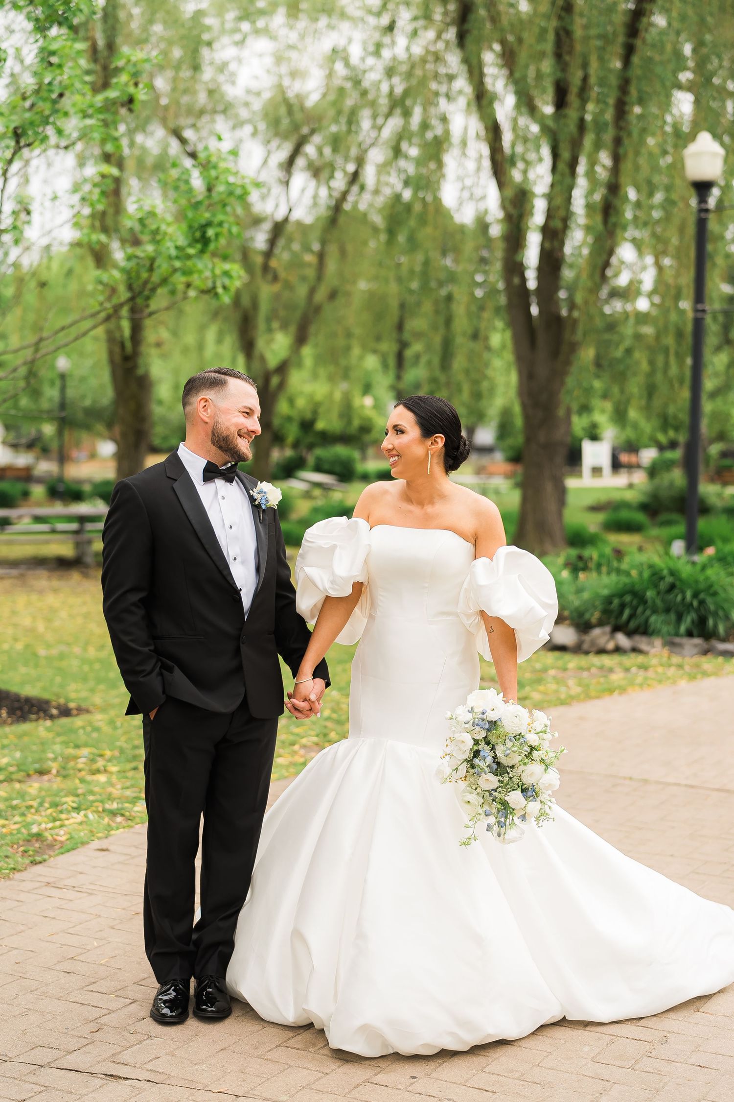 A bride in a flowing white gown with puff sleeves and groom in black tuxedo share a joyful moment on a garden path.