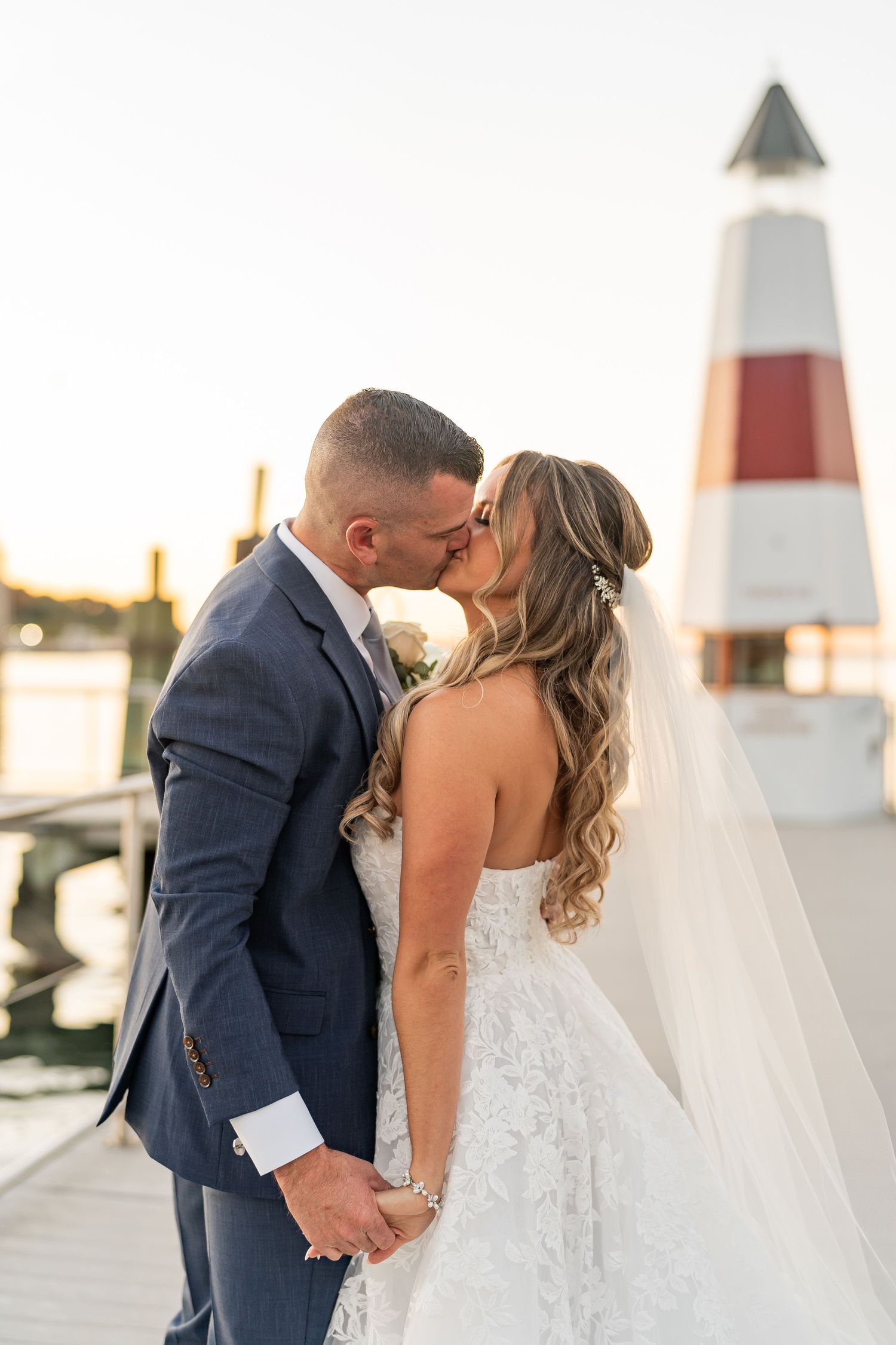 A romantic wedding couple shares a kiss at sunset near a lighthouse with warm golden lighting.
