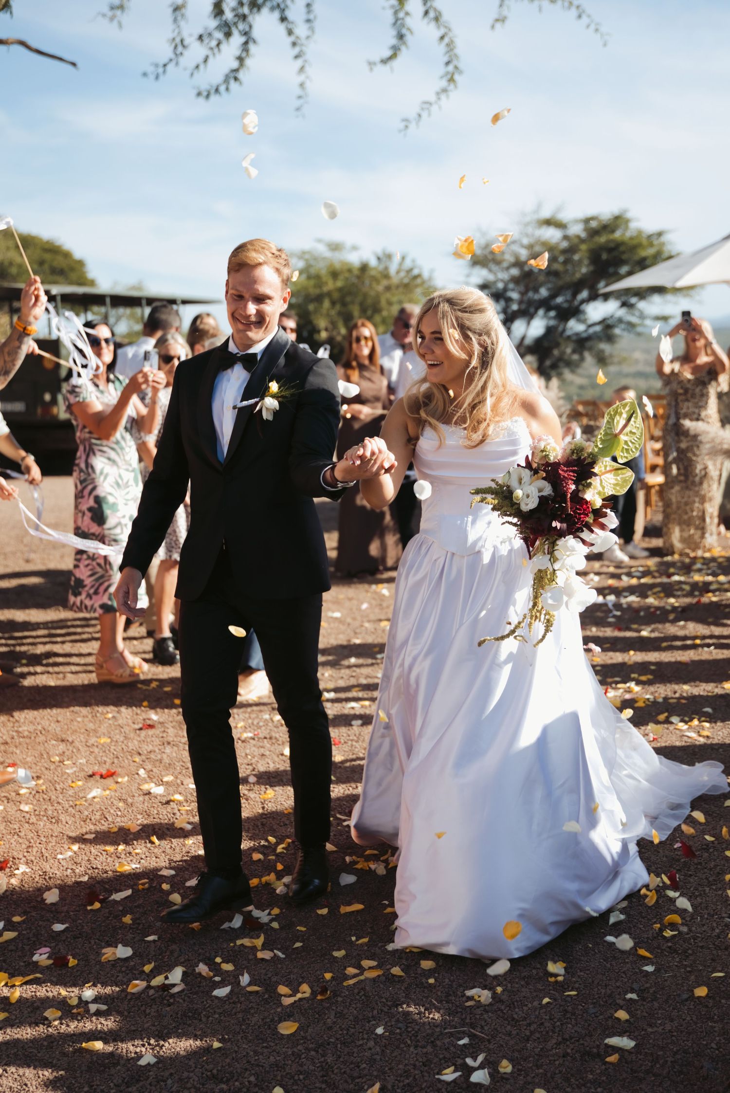 Bride and groom walk through flower petal confetti toss after outdoor wedding ceremony, bride in white gown holding bouquet.