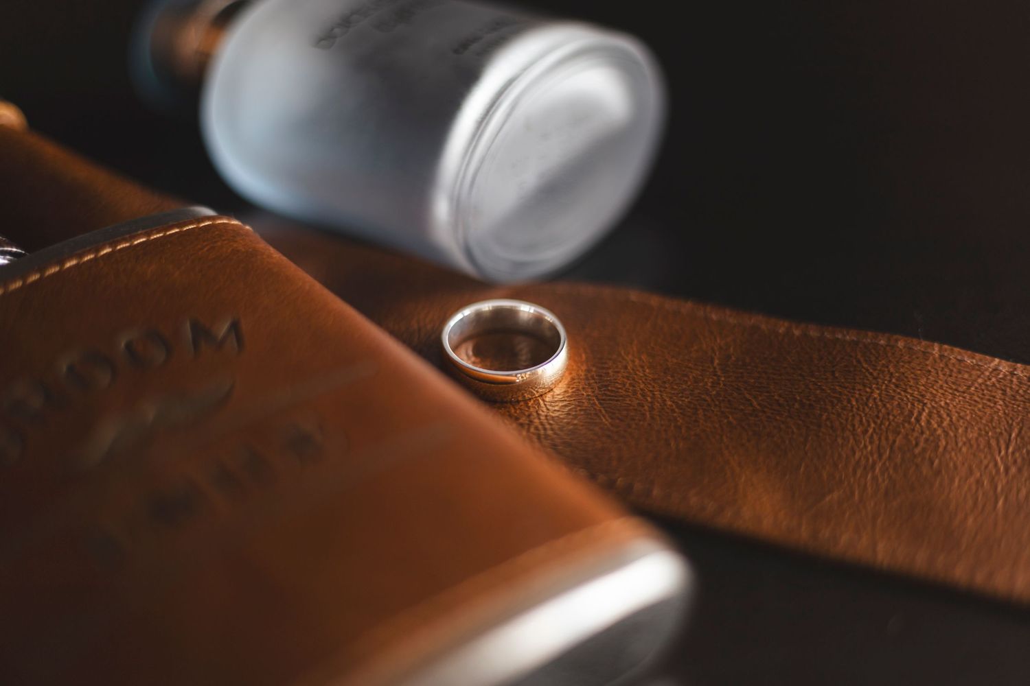 Wedding ring photographed on brown leather with flask in dark ambient lighting.