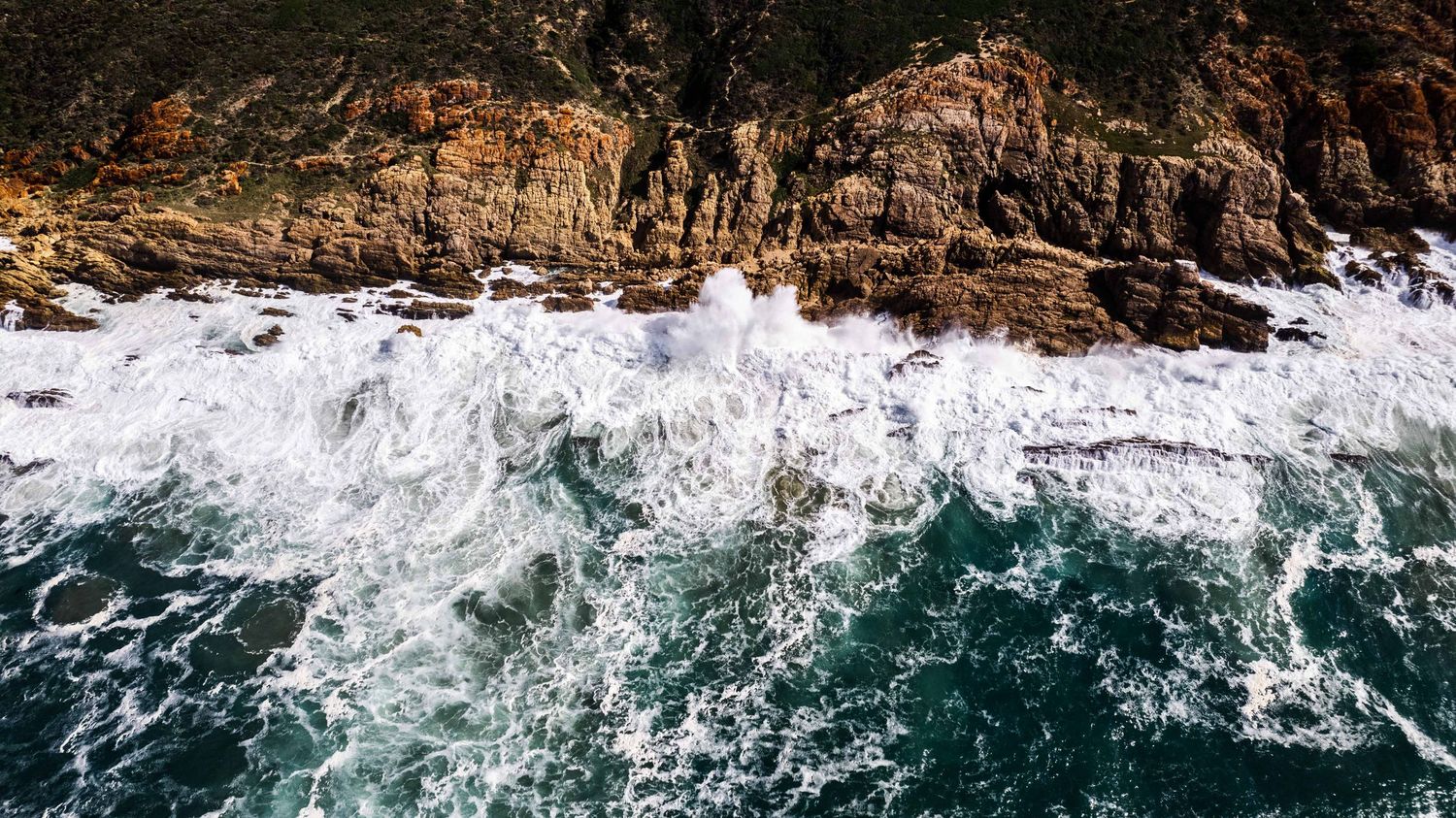 Dramatic aerial shot of powerful waves crashing against rocky coastal cliffs with turquoise water.