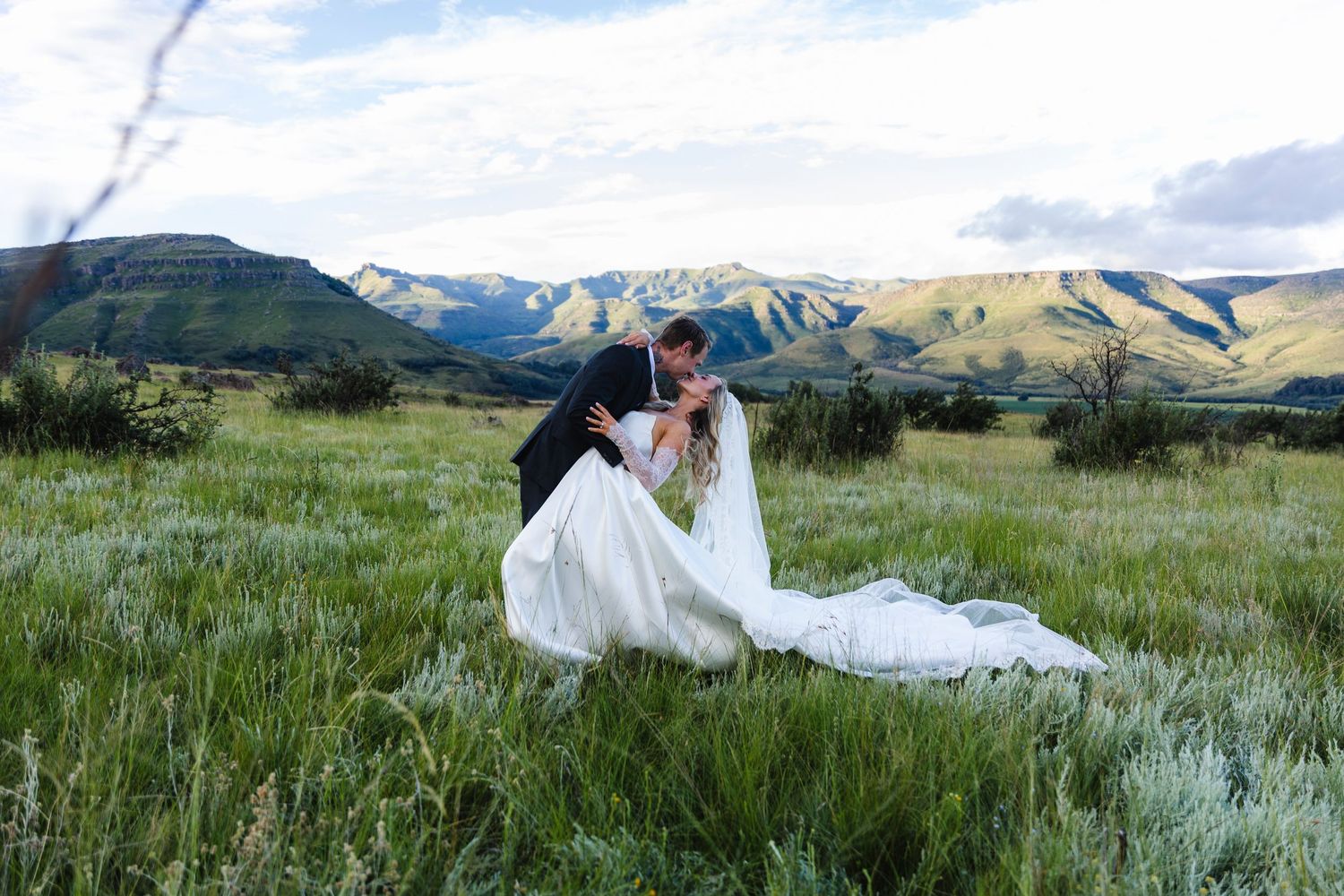 A bride and groom share a romantic dip kiss in a lush green meadow with stunning mountain views.