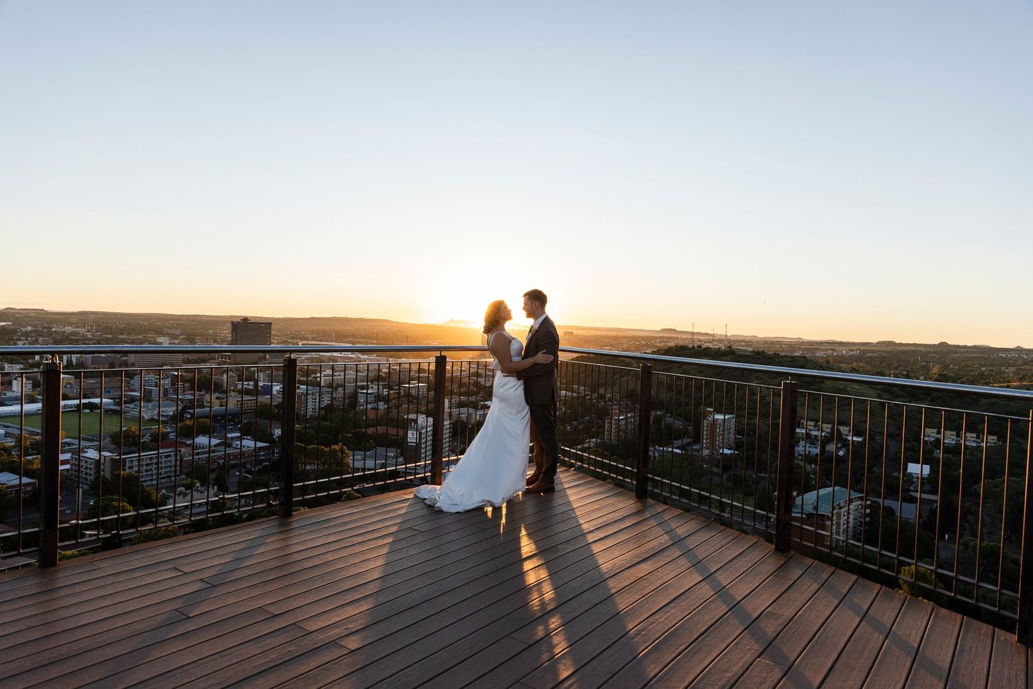 Bride and groom embrace on a rooftop deck at golden hour sunset with a panoramic city view behind them.