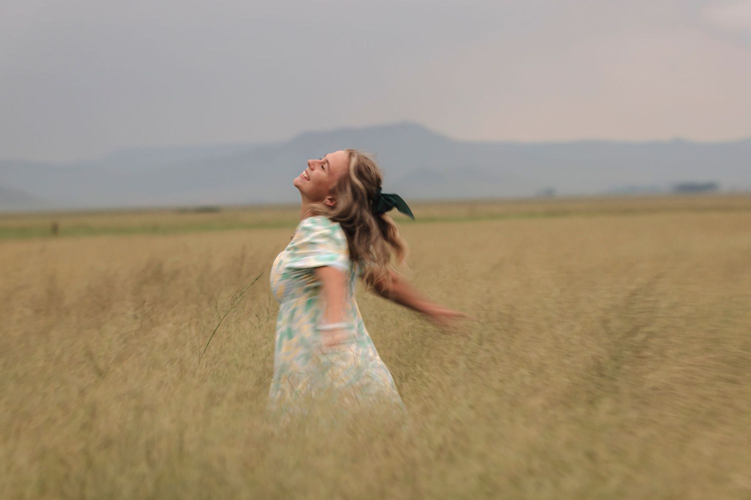 Woman in floral dress twirling joyfully in a golden field with mountains in background.