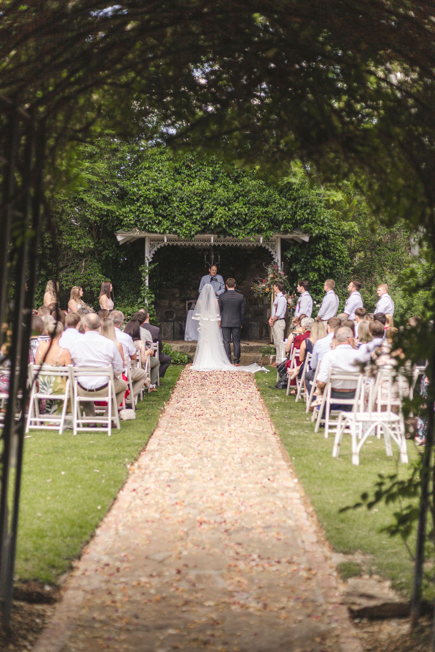 Garden wedding ceremony under pergola with flower-lined aisle and white chairs.