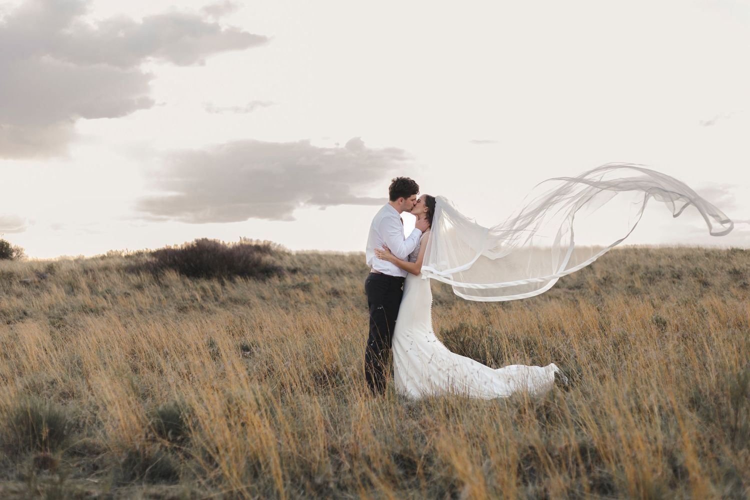 Wedding couple standing in field with bride's veil flowing dramatically in wind.