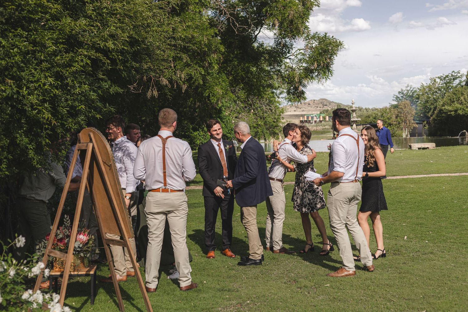 Wedding guests gather around an easel sign on manicured lawn during outdoor reception.