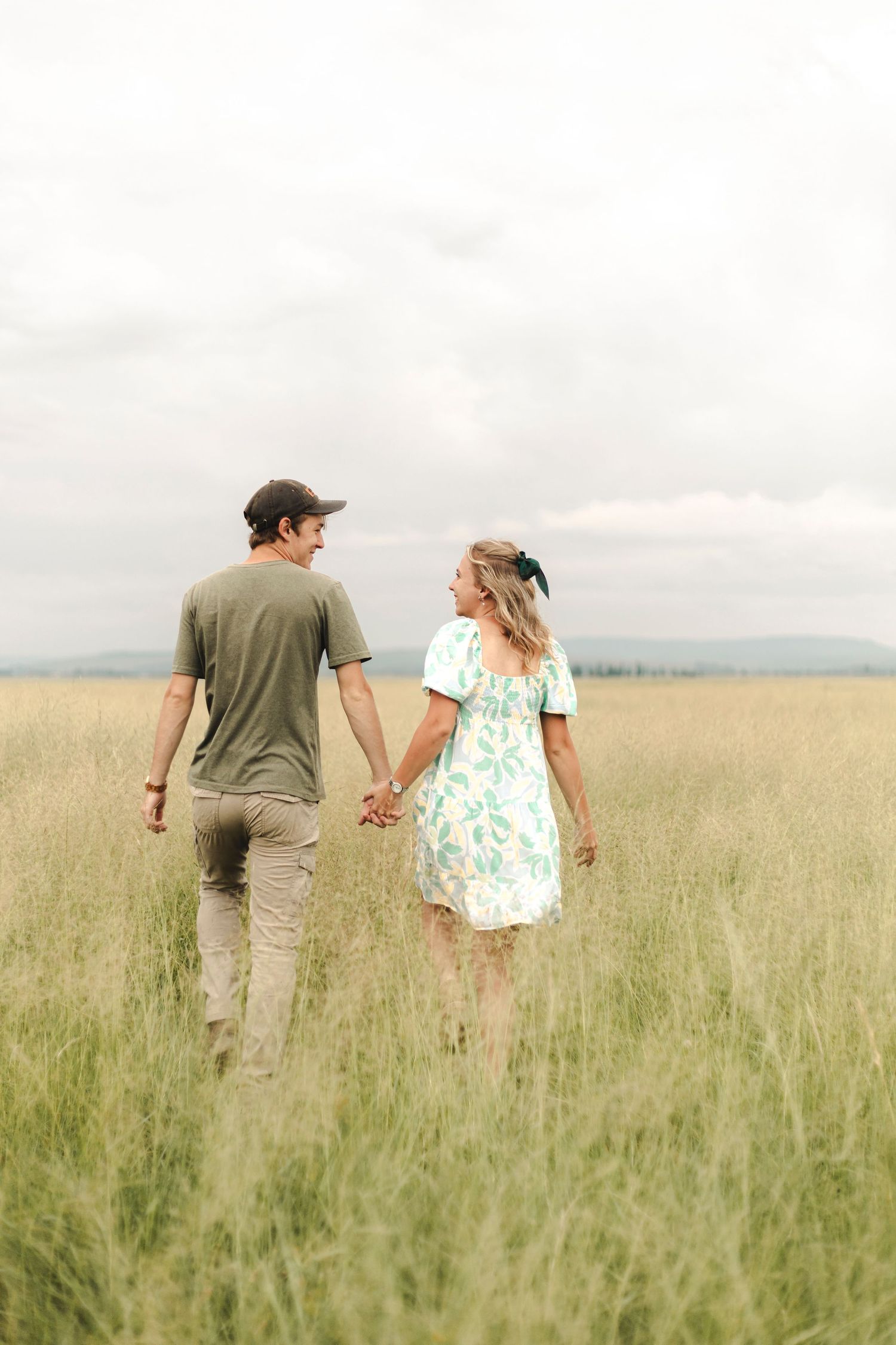 Couple walking hand in hand through golden field with mountains in background.