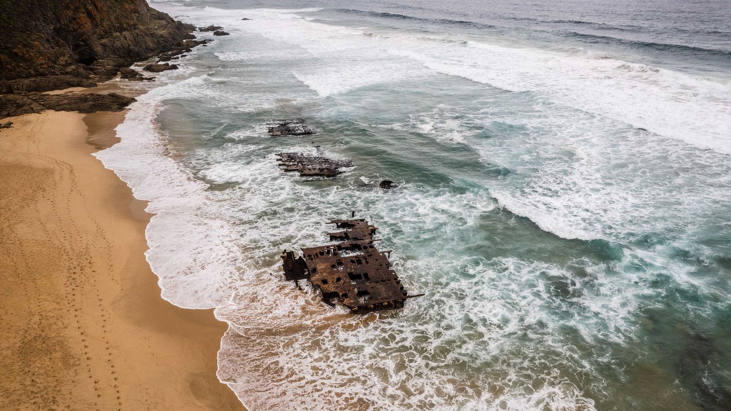 Aerial view of rugged coastline with turquoise waves crashing against sandy beach and rocky cliffs.