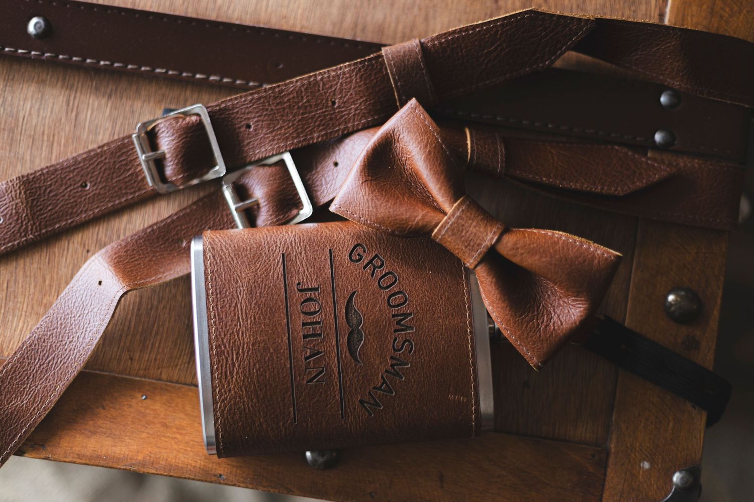 A leather hip flask and brown bow tie arranged on a wooden surface with leather straps.