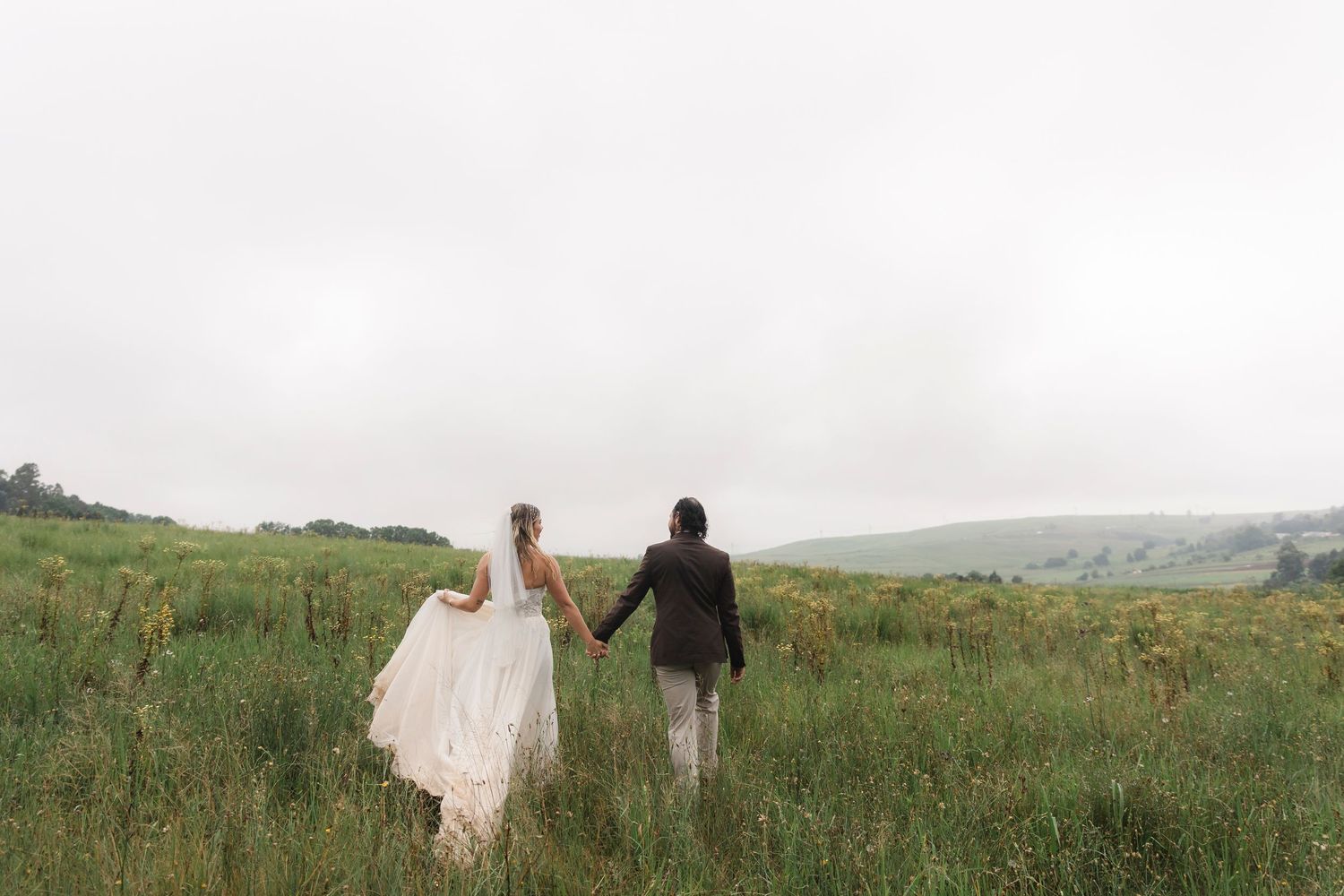 A bride and groom hold hands walking through a lush green field on their wedding day.
