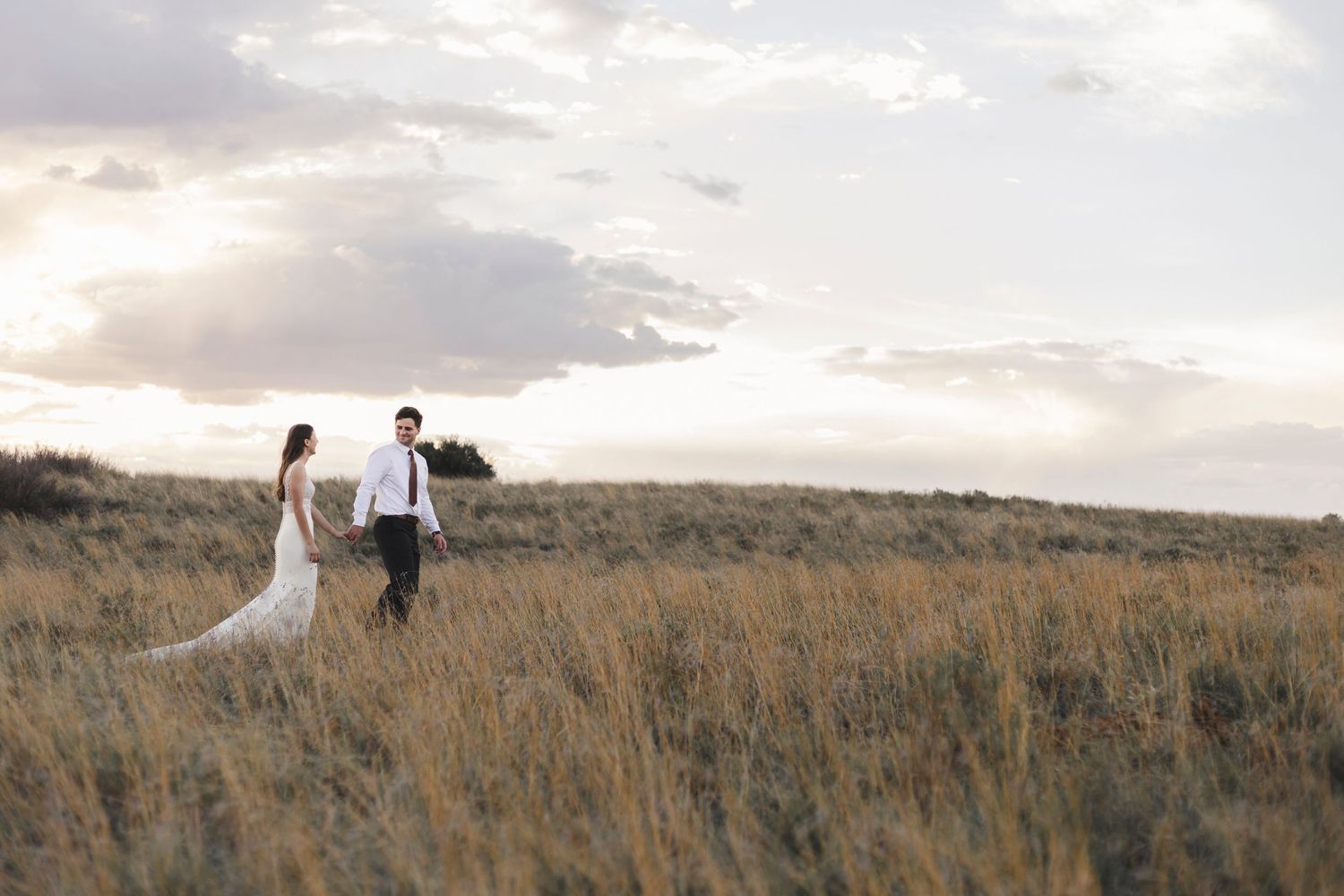 A couple walking hand in hand through a golden prairie field at sunset.