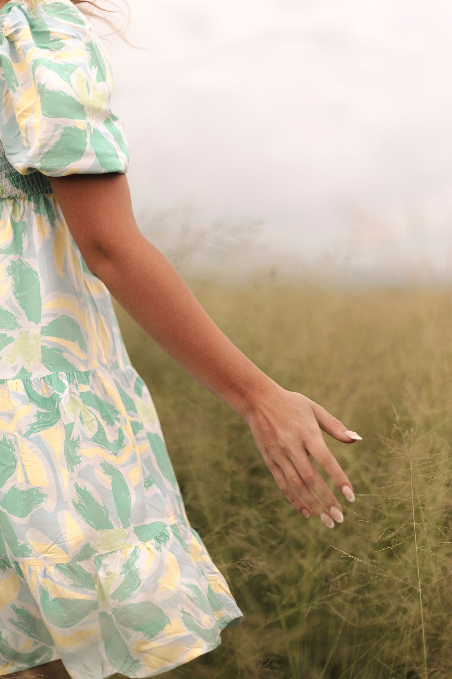 Woman in floral dress running hand through tall grass in a serene meadow.