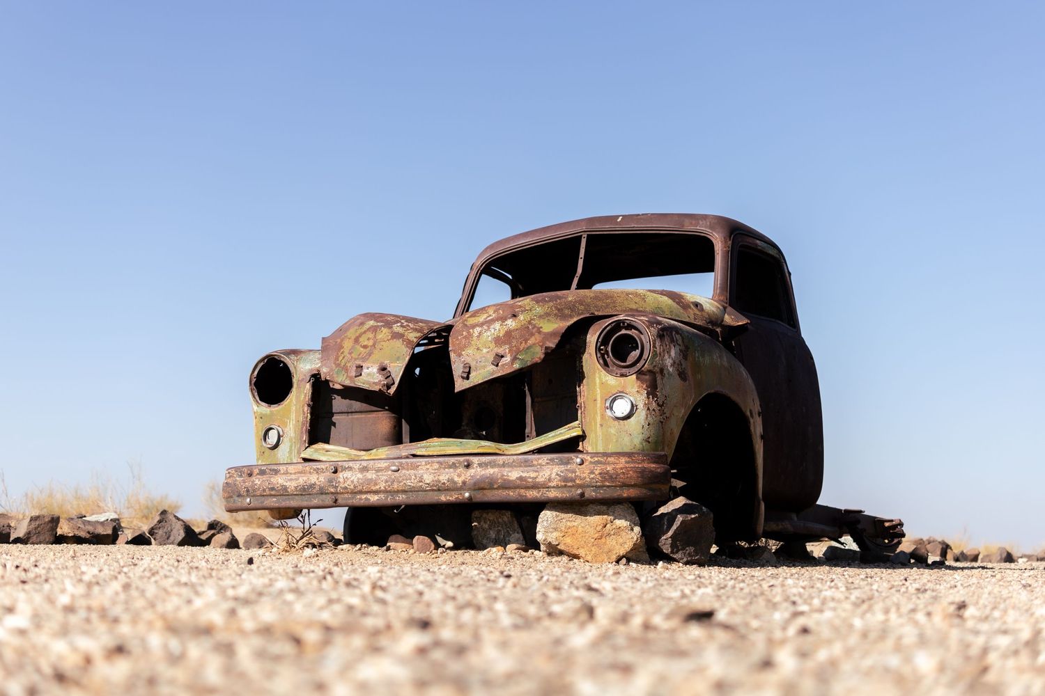 Rusted vintage car wreck abandoned in desert landscape against clear blue sky.