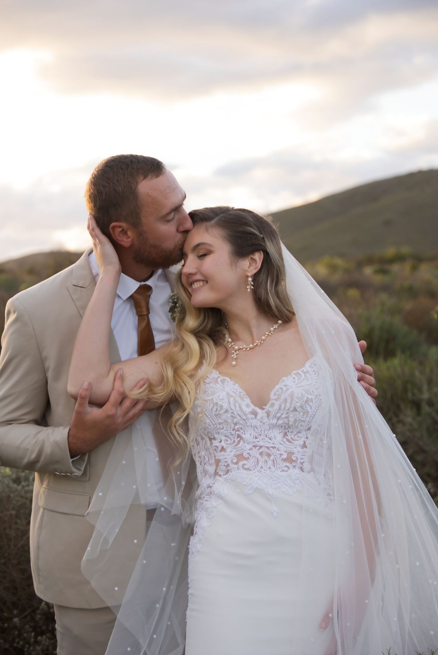 A groom in a tan suit kisses his smiling bride in a lace wedding dress and veil outdoors at sunset.