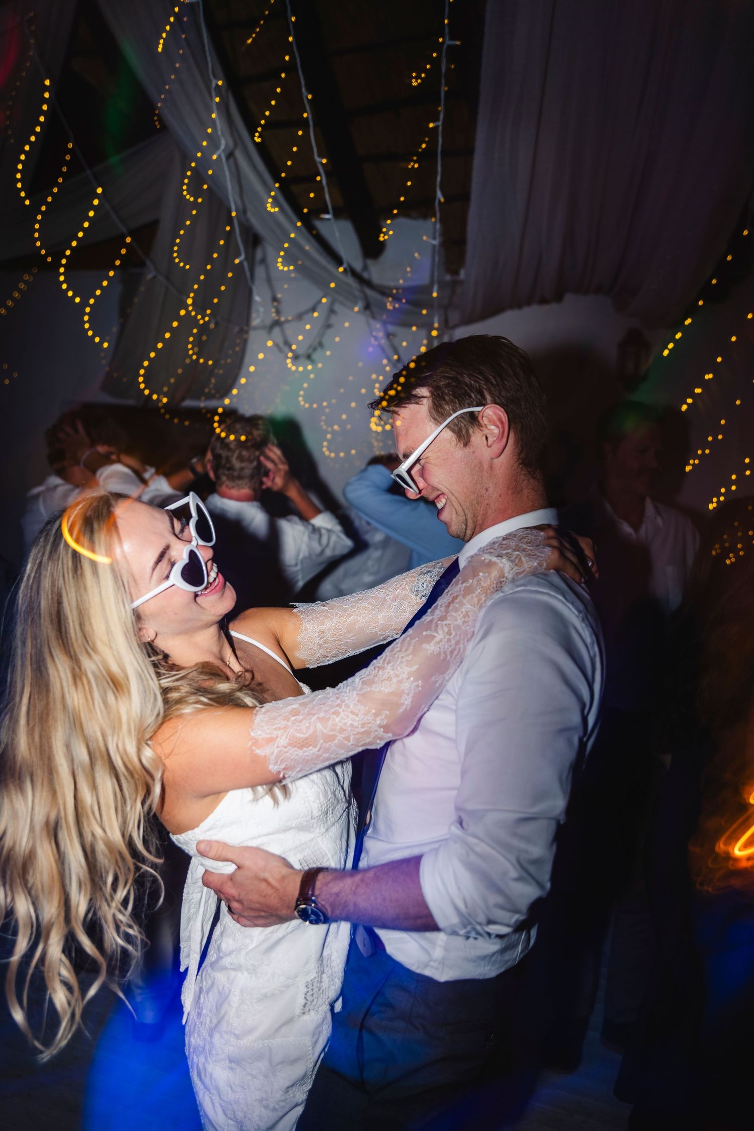 A bride and groom dancing joyfully wearing white novelty sunglasses at their wedding reception with string lights in background.