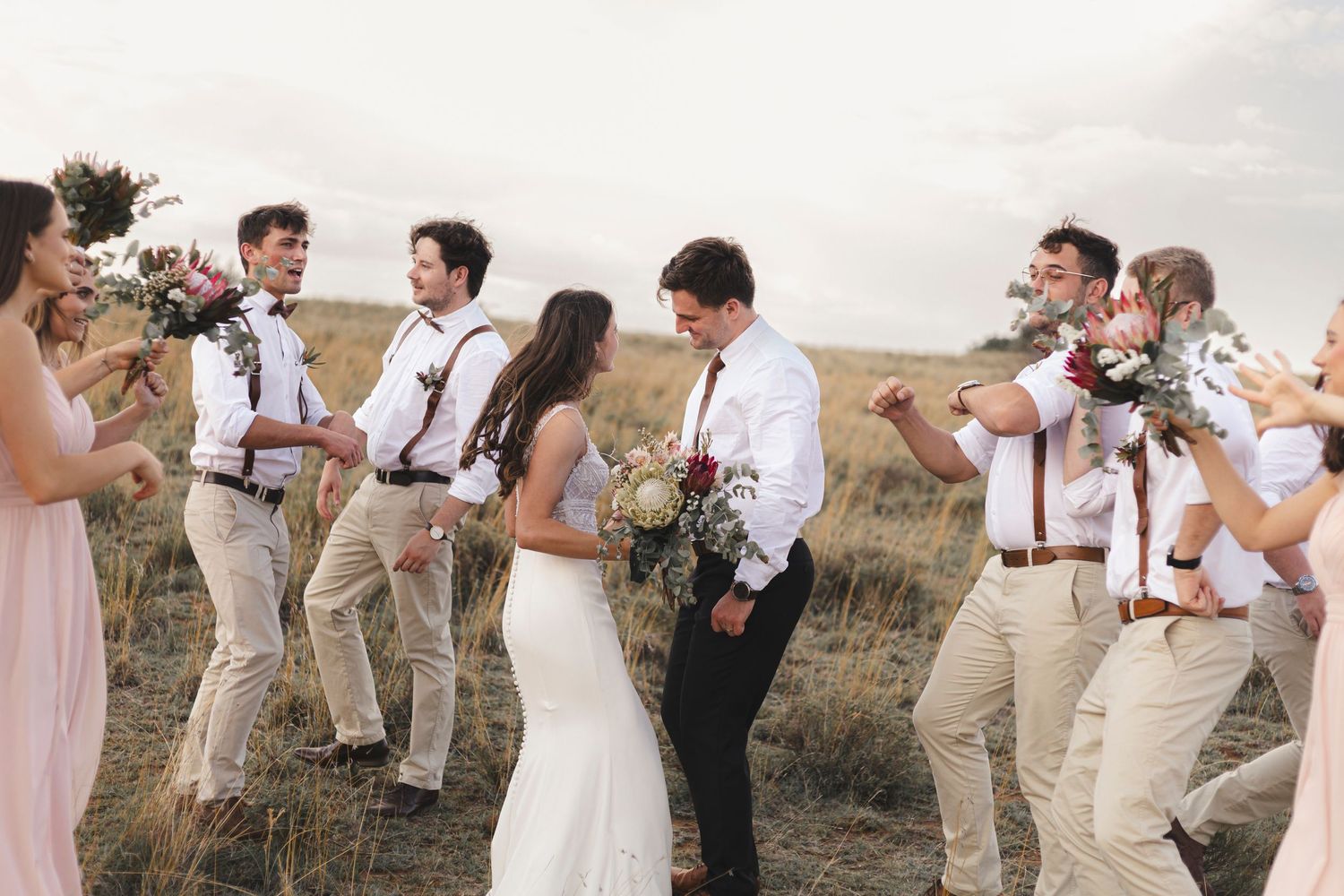 Wedding party celebrating in field with groomsmen in khaki pants and suspenders.