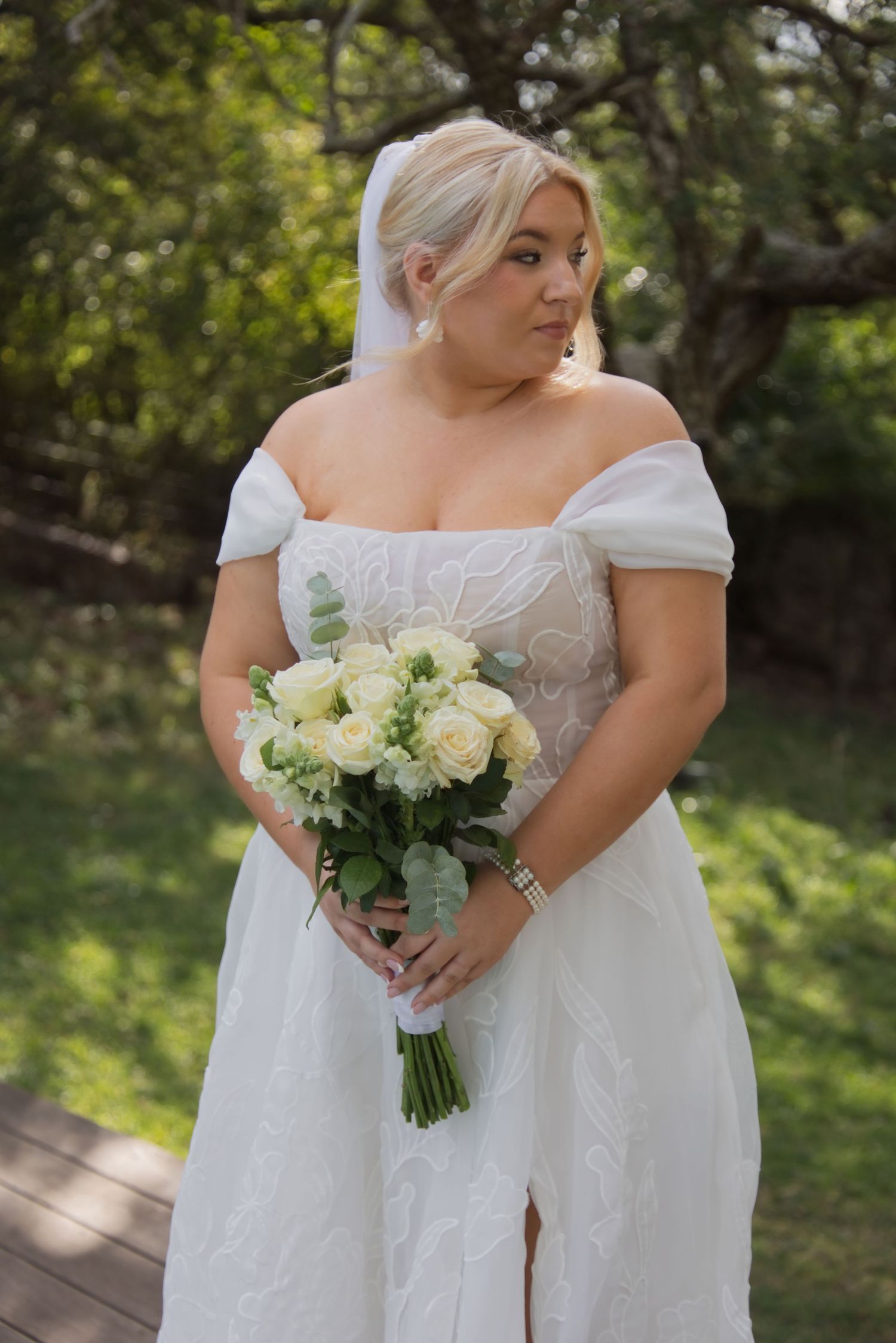 Bride in off-shoulder white lace gown holds ivory rose bouquet outdoors, with veil and elegant jewelry.