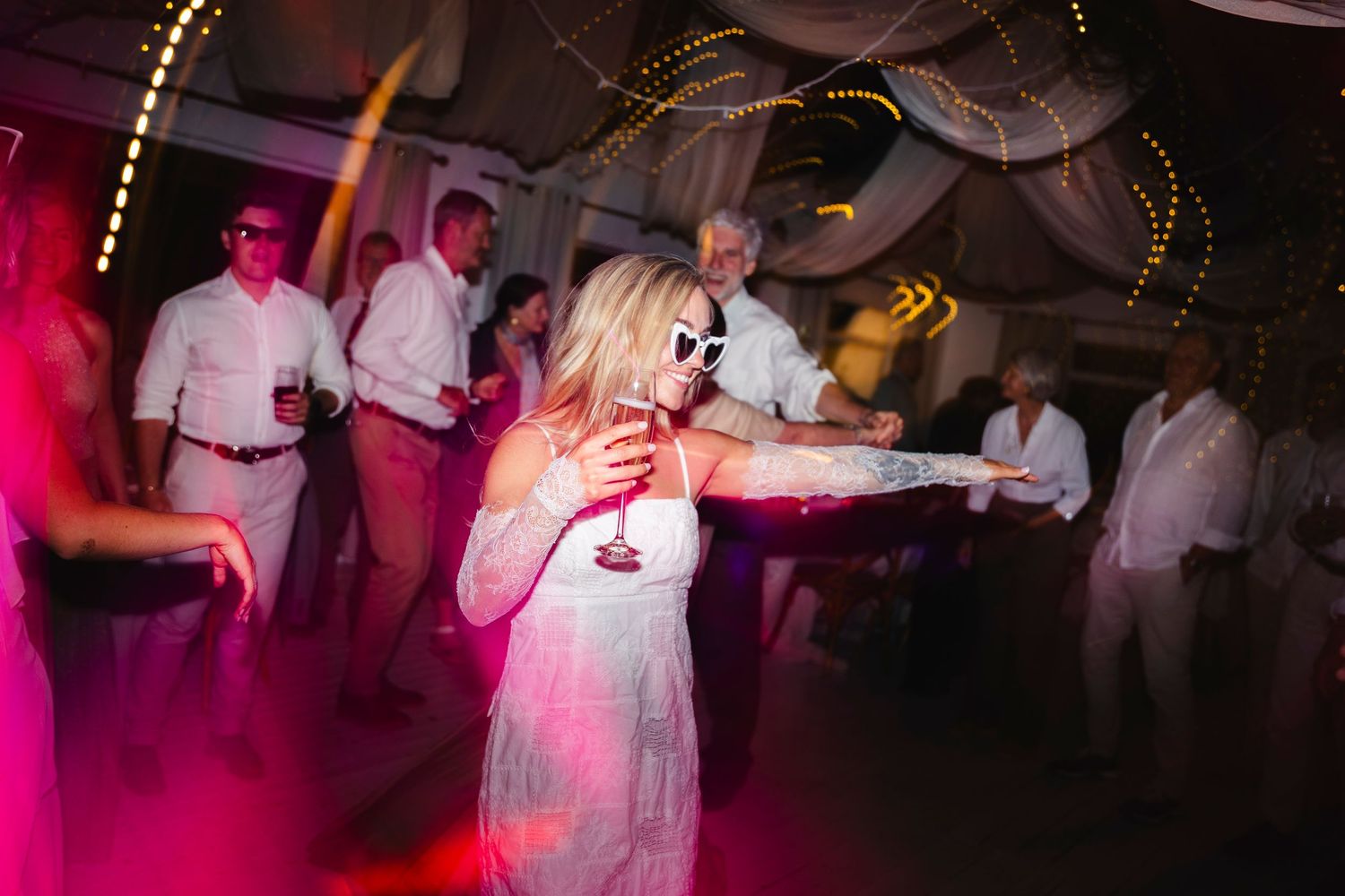 Woman in white dress and sunglasses dancing at a lively wedding reception under pink lights.