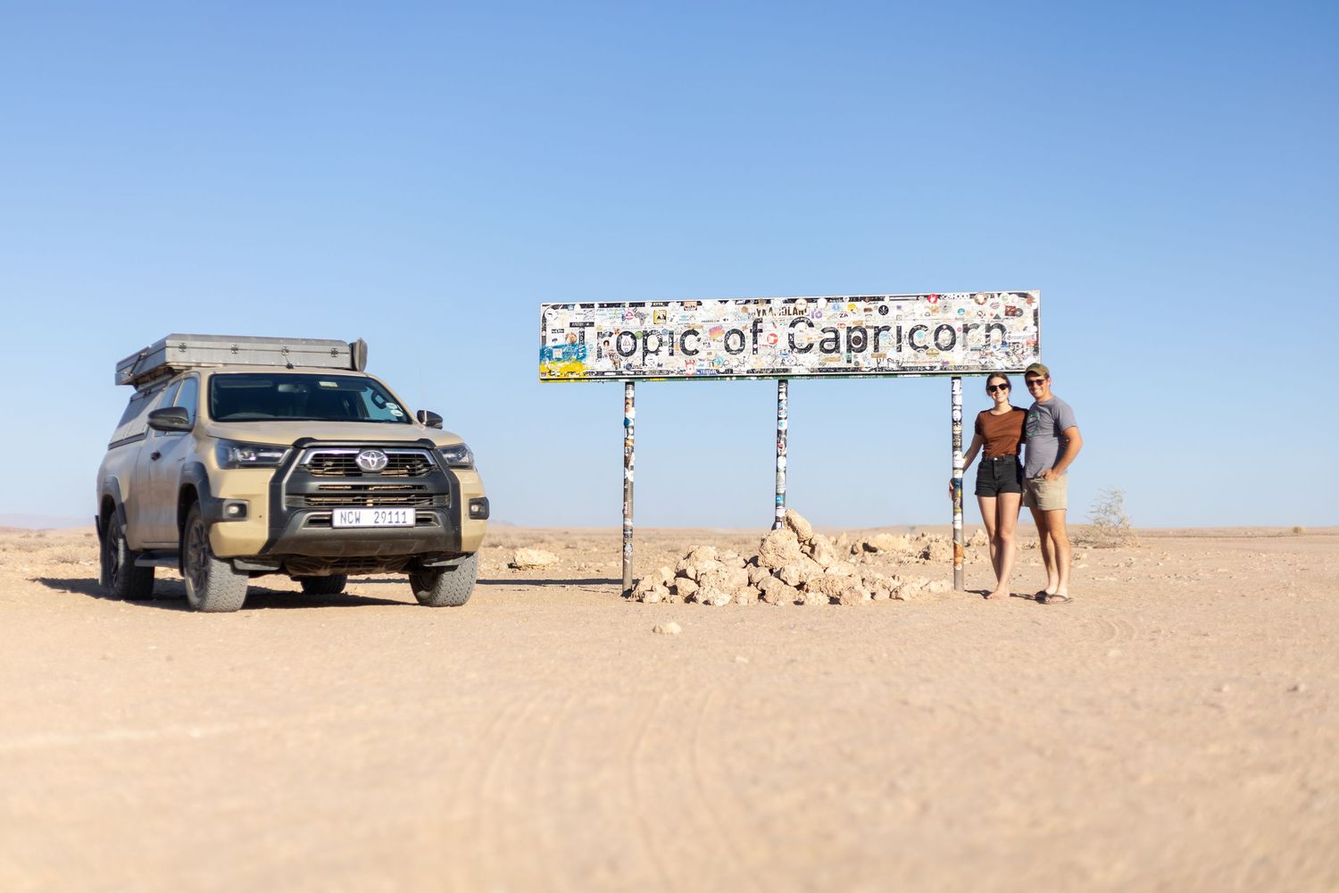 An SUV and travelers stand by the Tropic of Capricorn marker in a desert landscape.