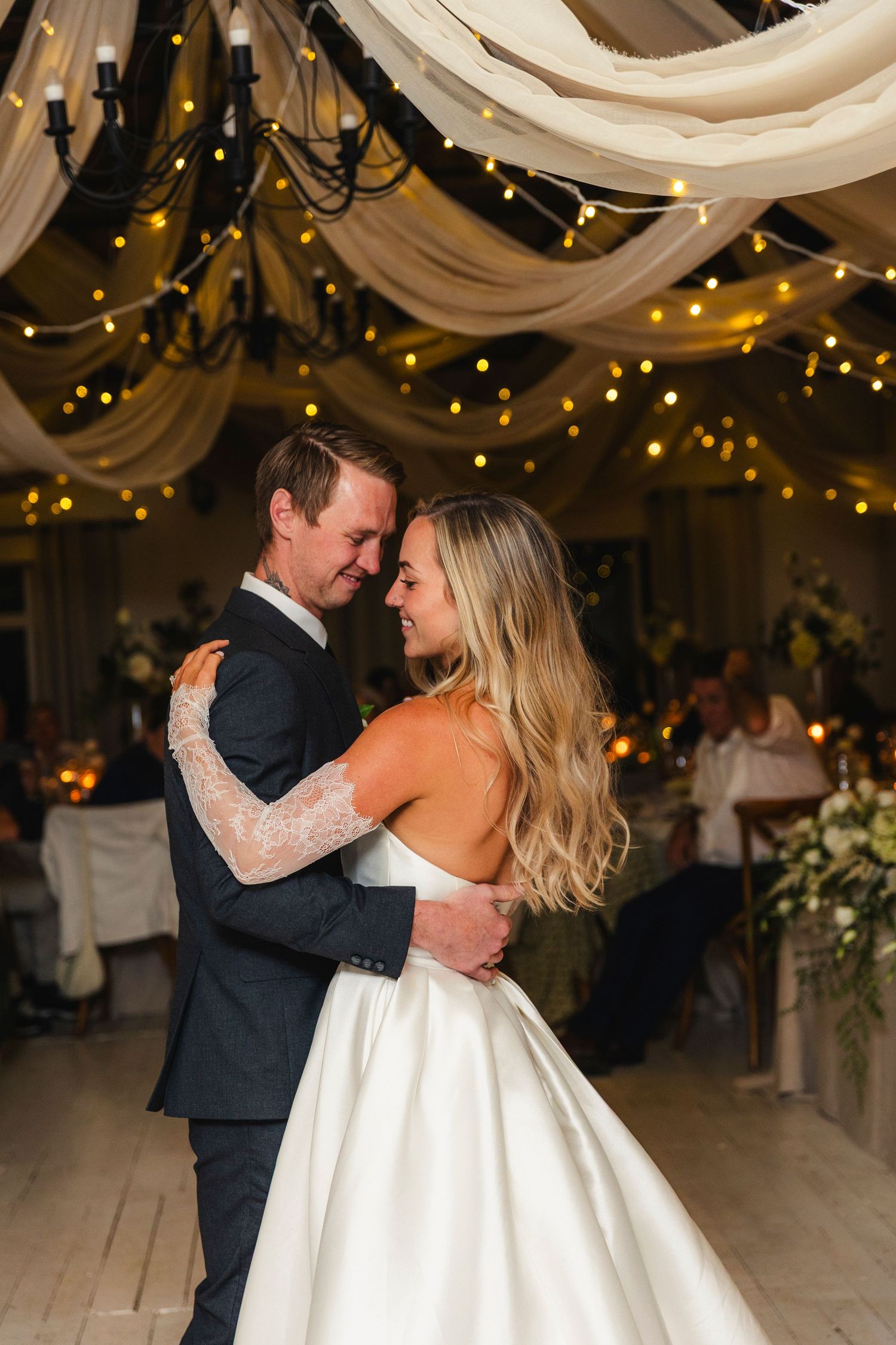 Bride and groom share their first dance under fairy lights and draped fabric at elegant wedding reception.