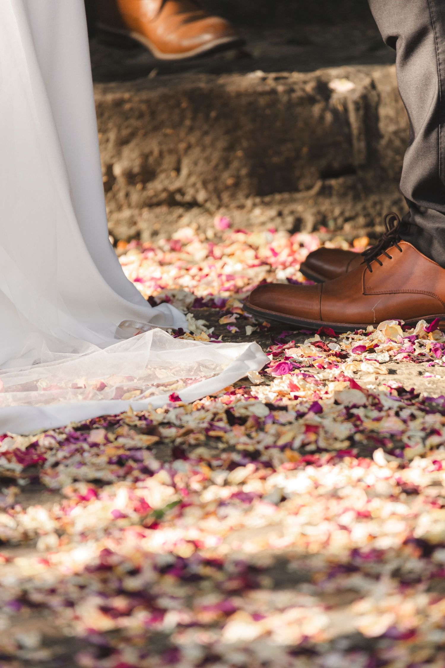 Brown leather dress shoes standing on scattered rose petals next to white fabric.
