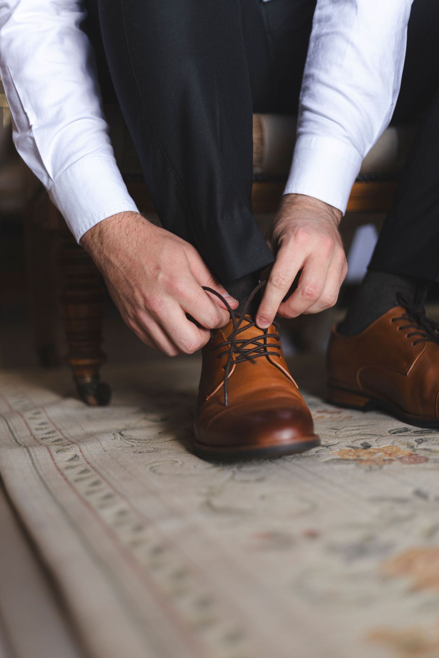 Close up of hands tying brown leather dress shoes on textured surface.