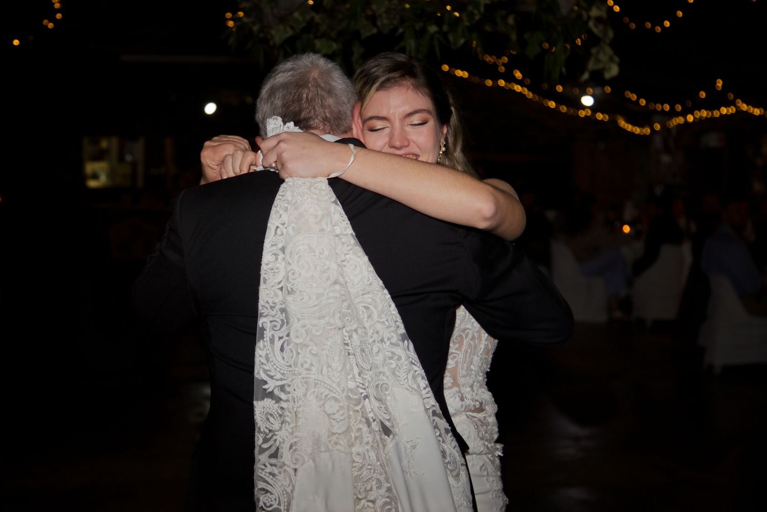 Bride in lace wedding gown shares an emotional hug with an older gentleman during an outdoor evening wedding reception.
