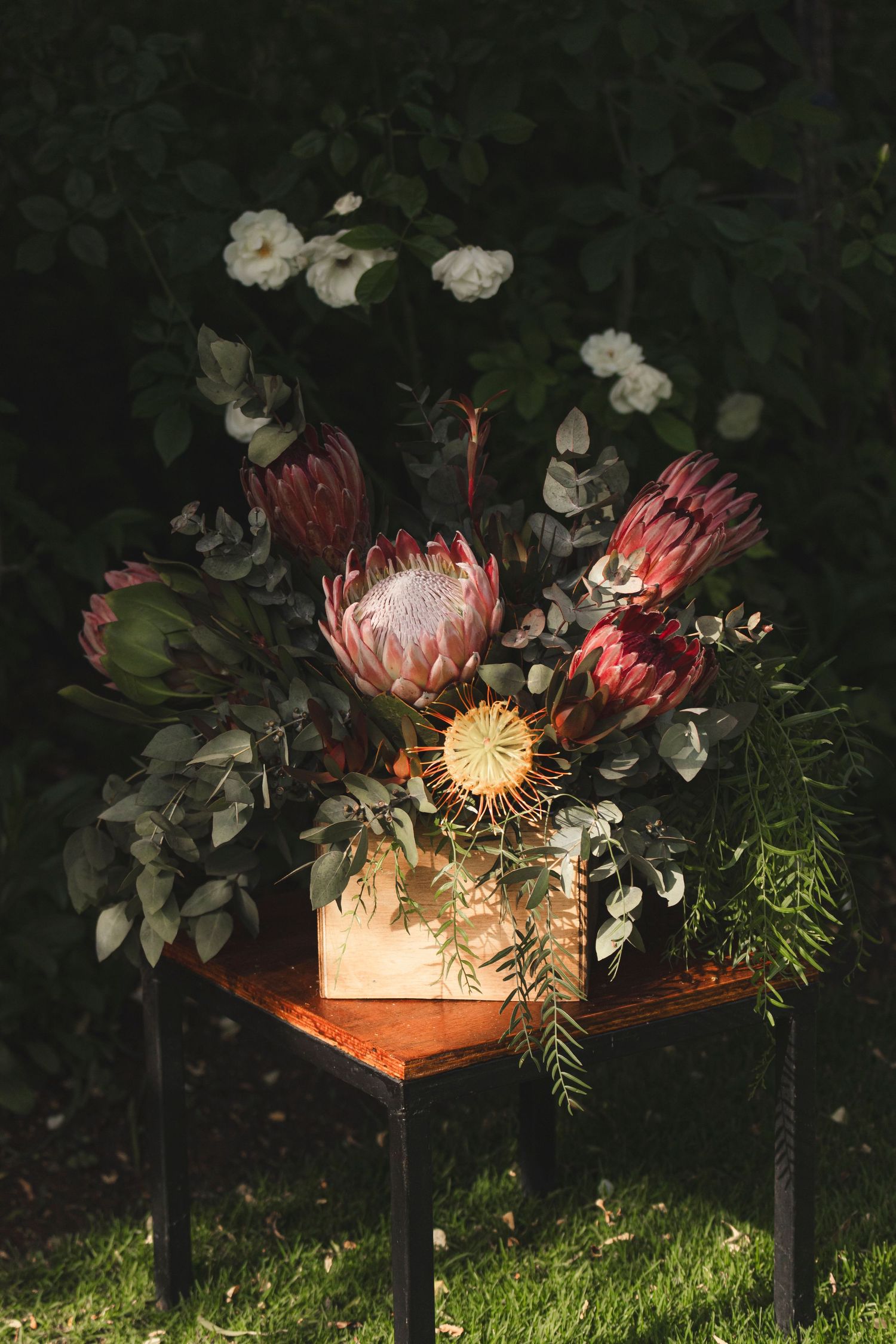 A dramatic floral arrangement with protea flowers and white blooms displayed on a dark wooden stand in a garden setting.