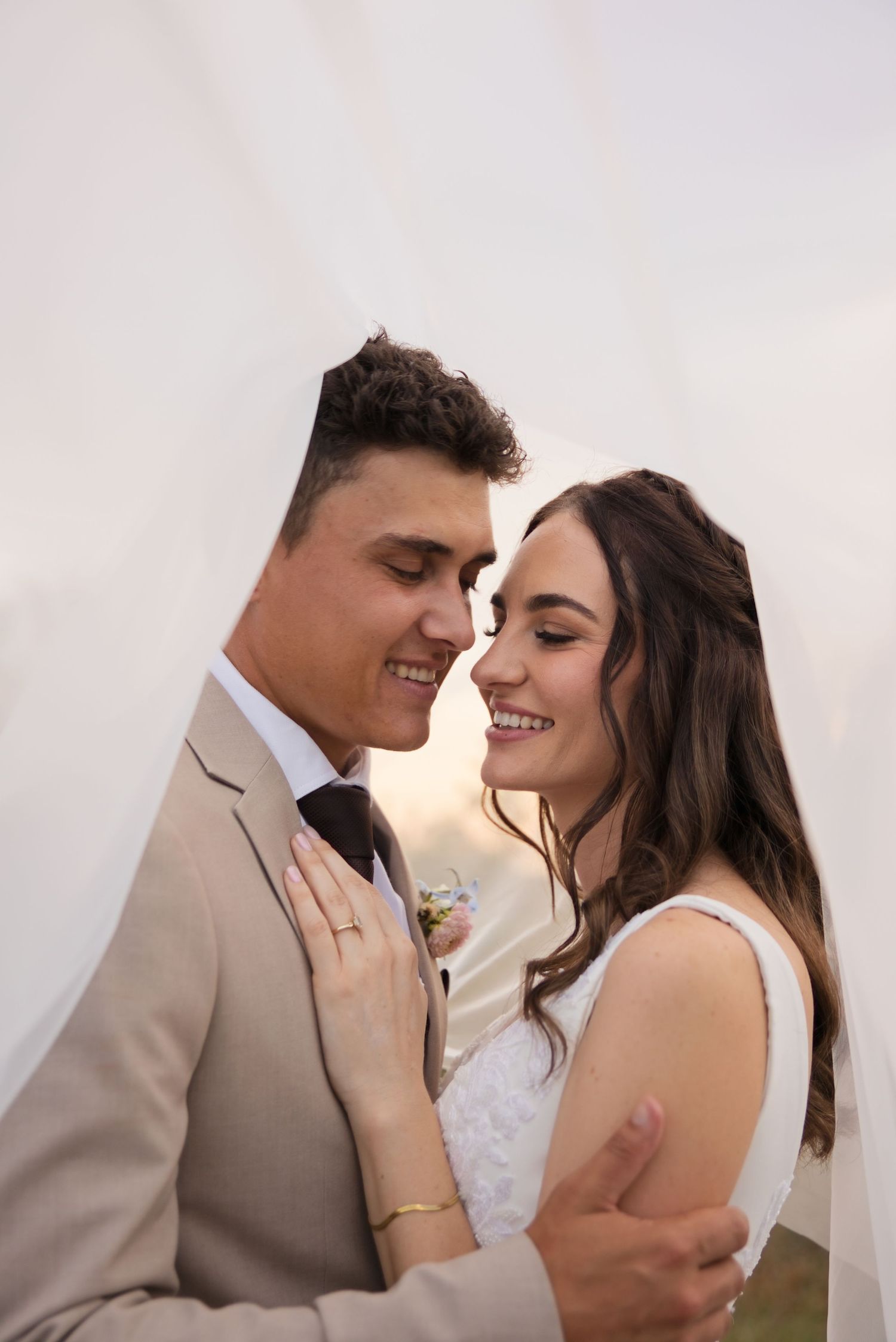 Bride and groom share a romantic moment, smiling closely together, groom in tan suit, bride in white lace dress.