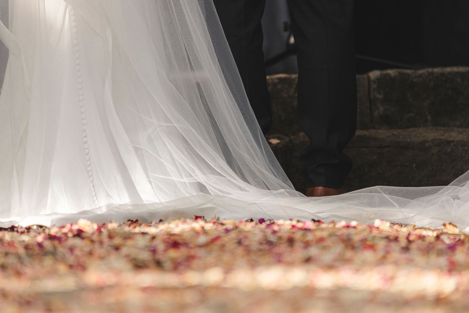 White wedding dress train flowing over scattered rose petals on stone ground.