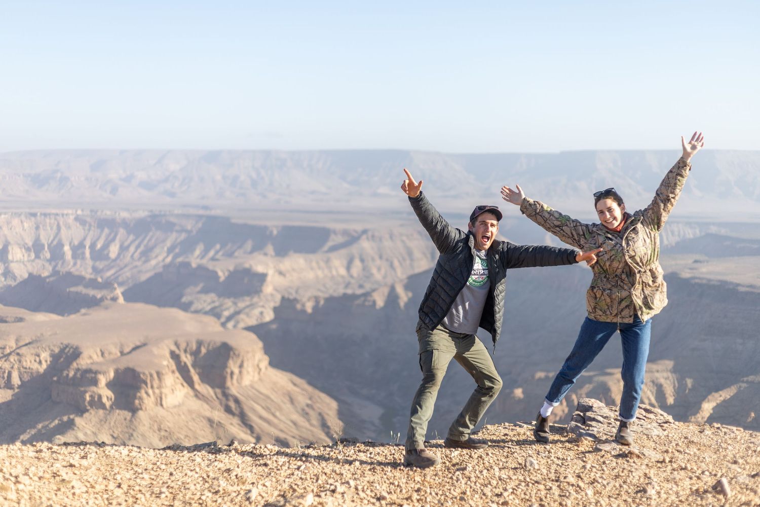 Two travelers pose energetically on the rim of a rocky desert canyon.