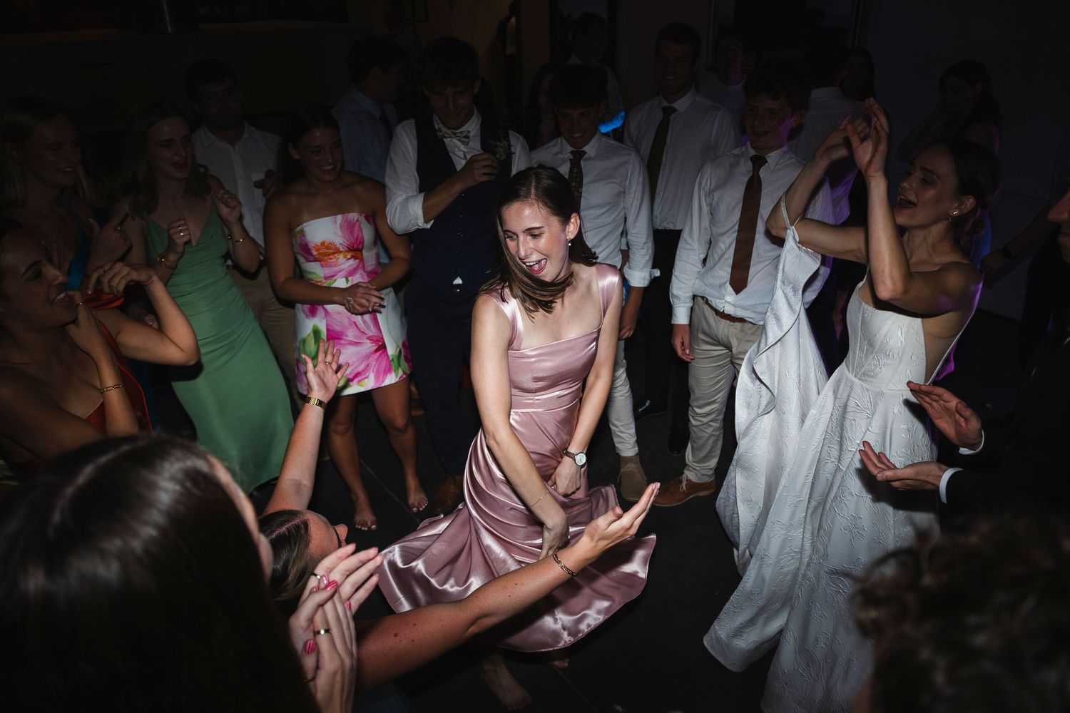 Guests dancing energetically at a wedding reception party, woman in pink satin dress at center of crowded dance floor.