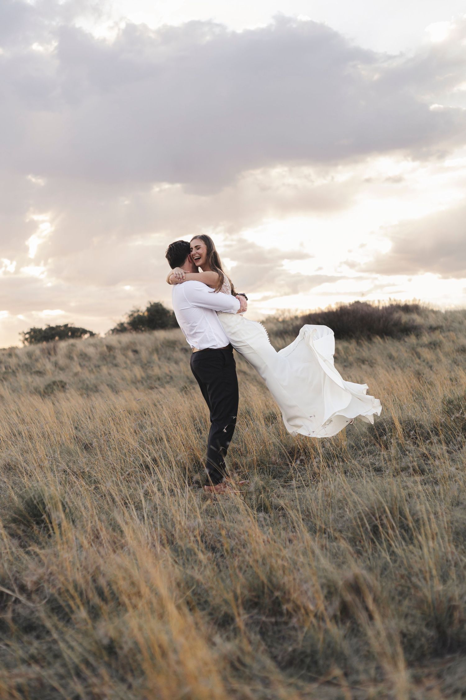 Groom lifting bride in white dress in golden field at sunset, sharing joyful embrace outdoors.