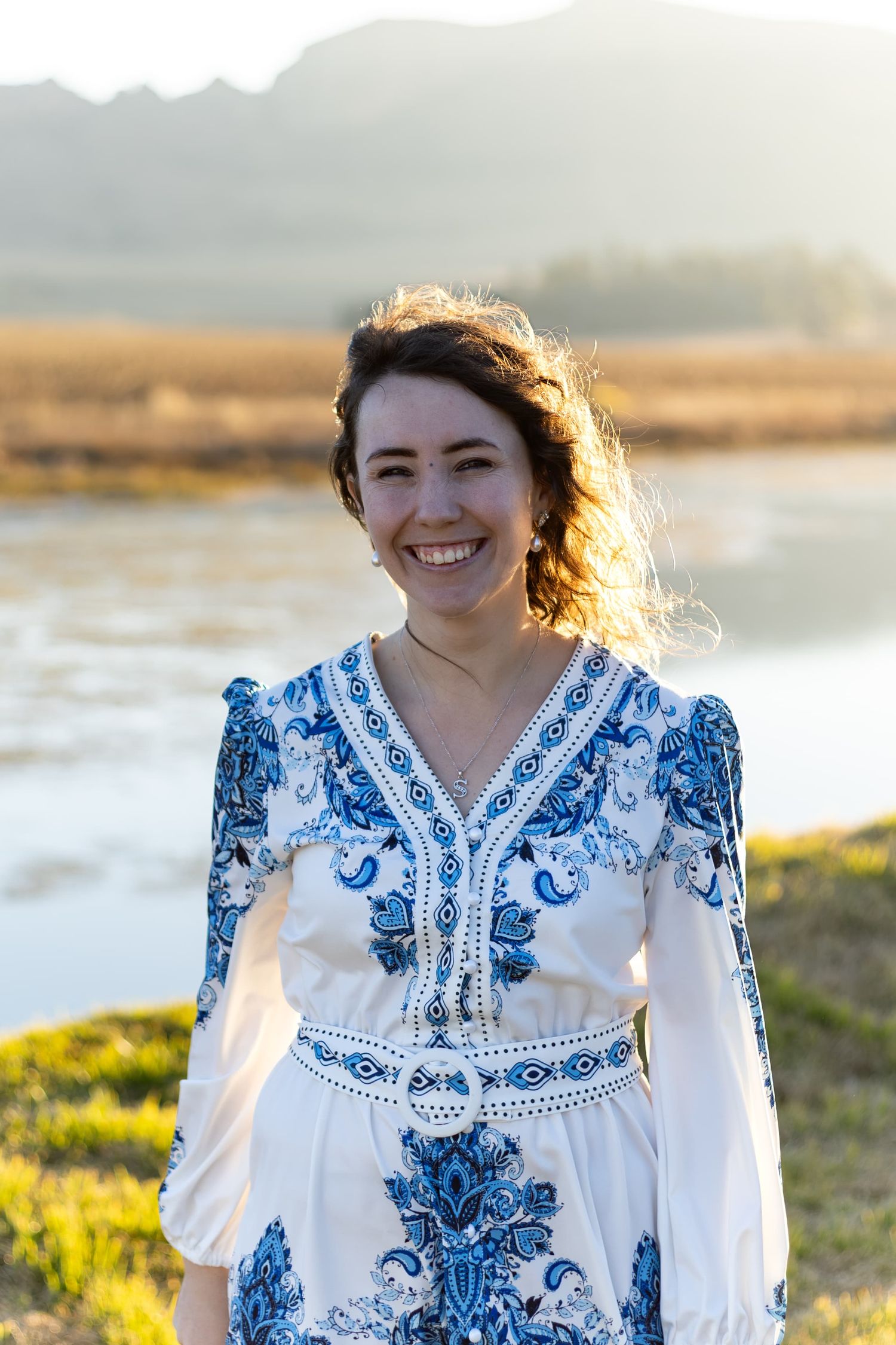 Smiling young woman in blue and white floral dress standing outdoors near a river with golden hills at sunset.