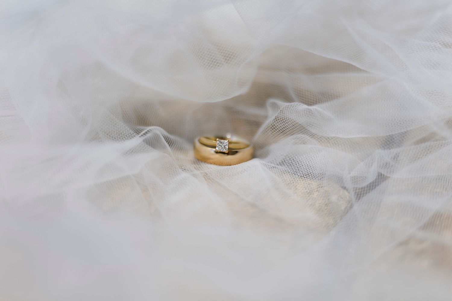 Gold wedding band and diamond engagement ring resting on white bridal veil fabric.