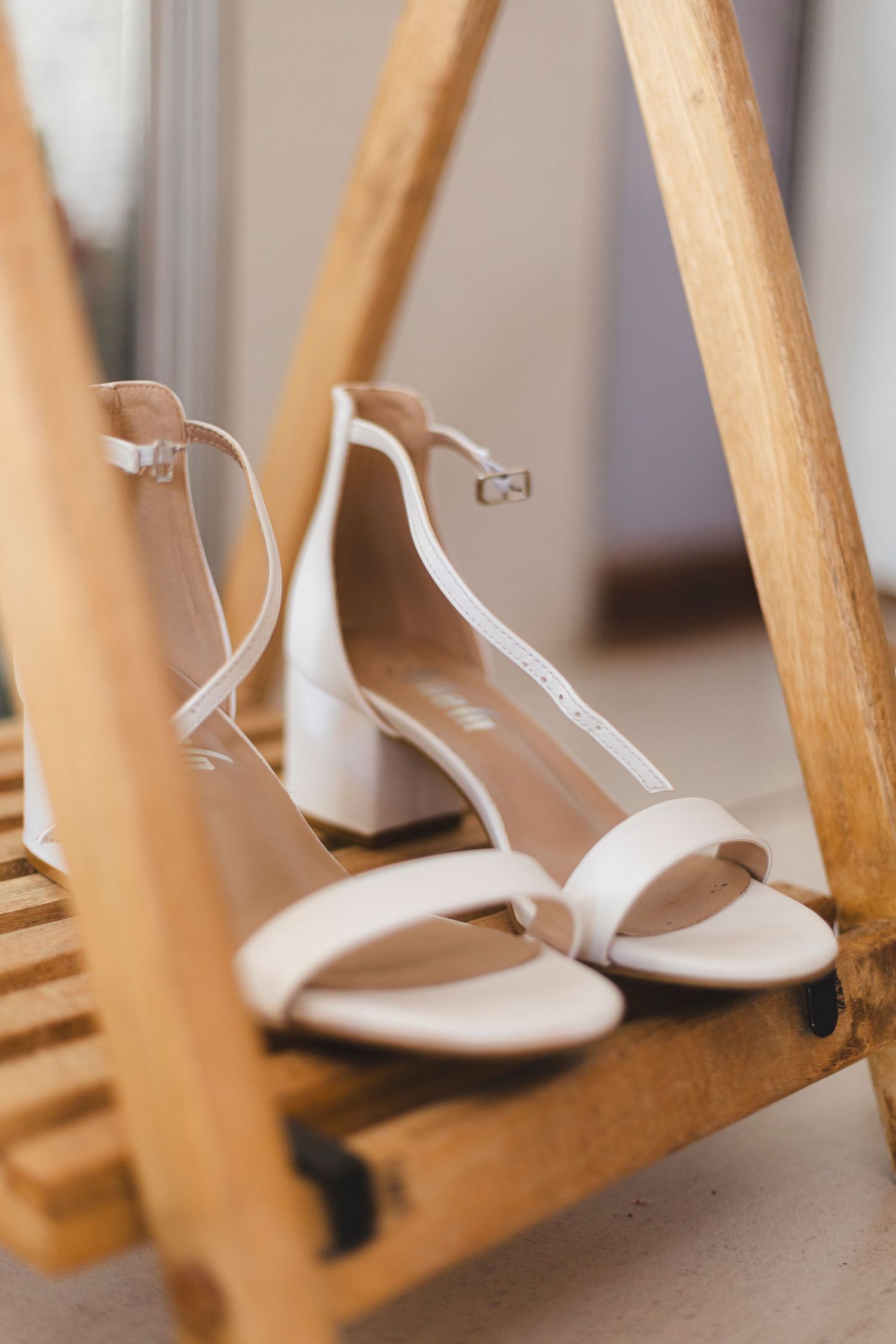 Elegant white bridal sandals with ankle straps displayed on wooden furniture.