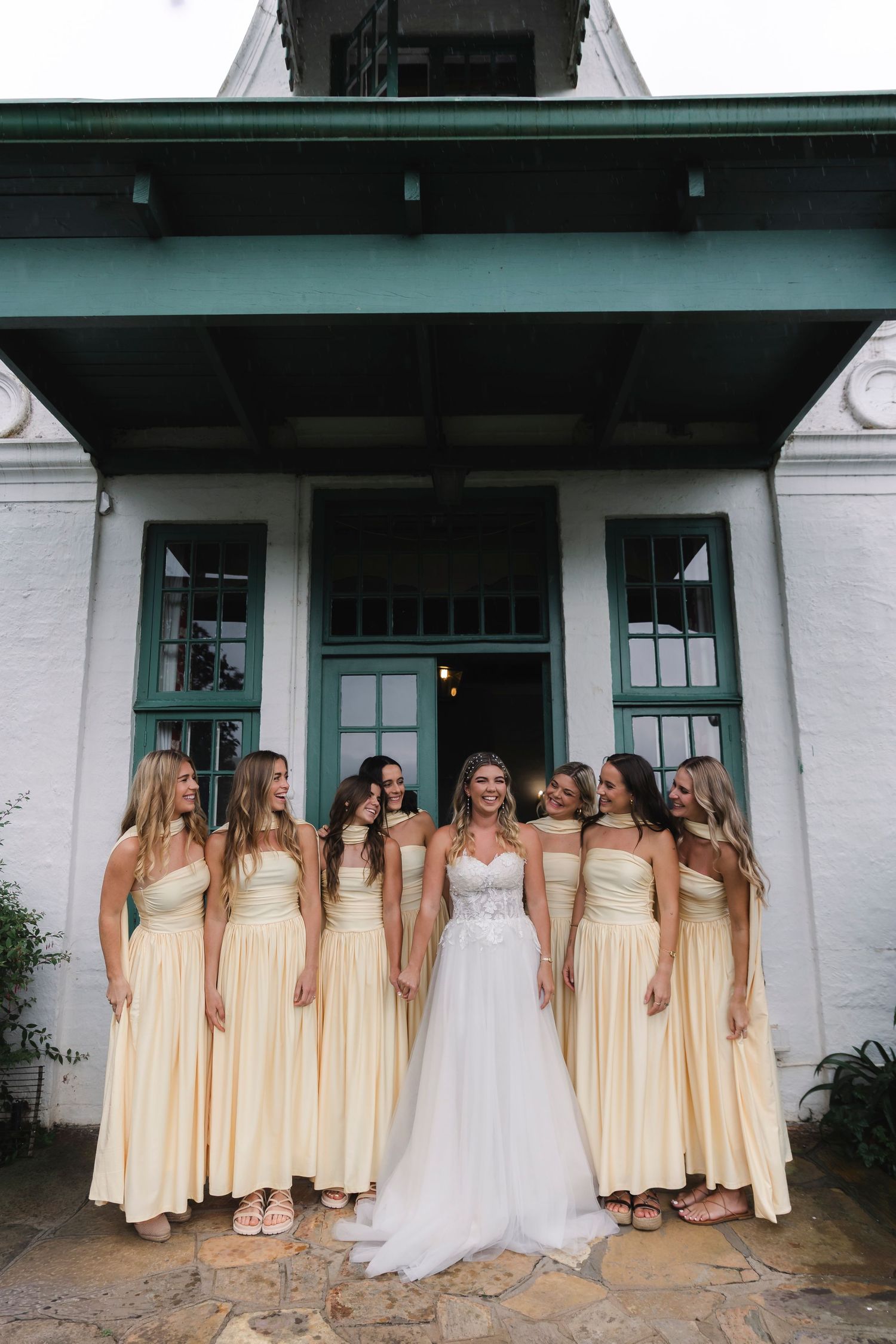 Bride in white gown poses with six bridesmaids in yellow dresses outside a white building with teal trim.