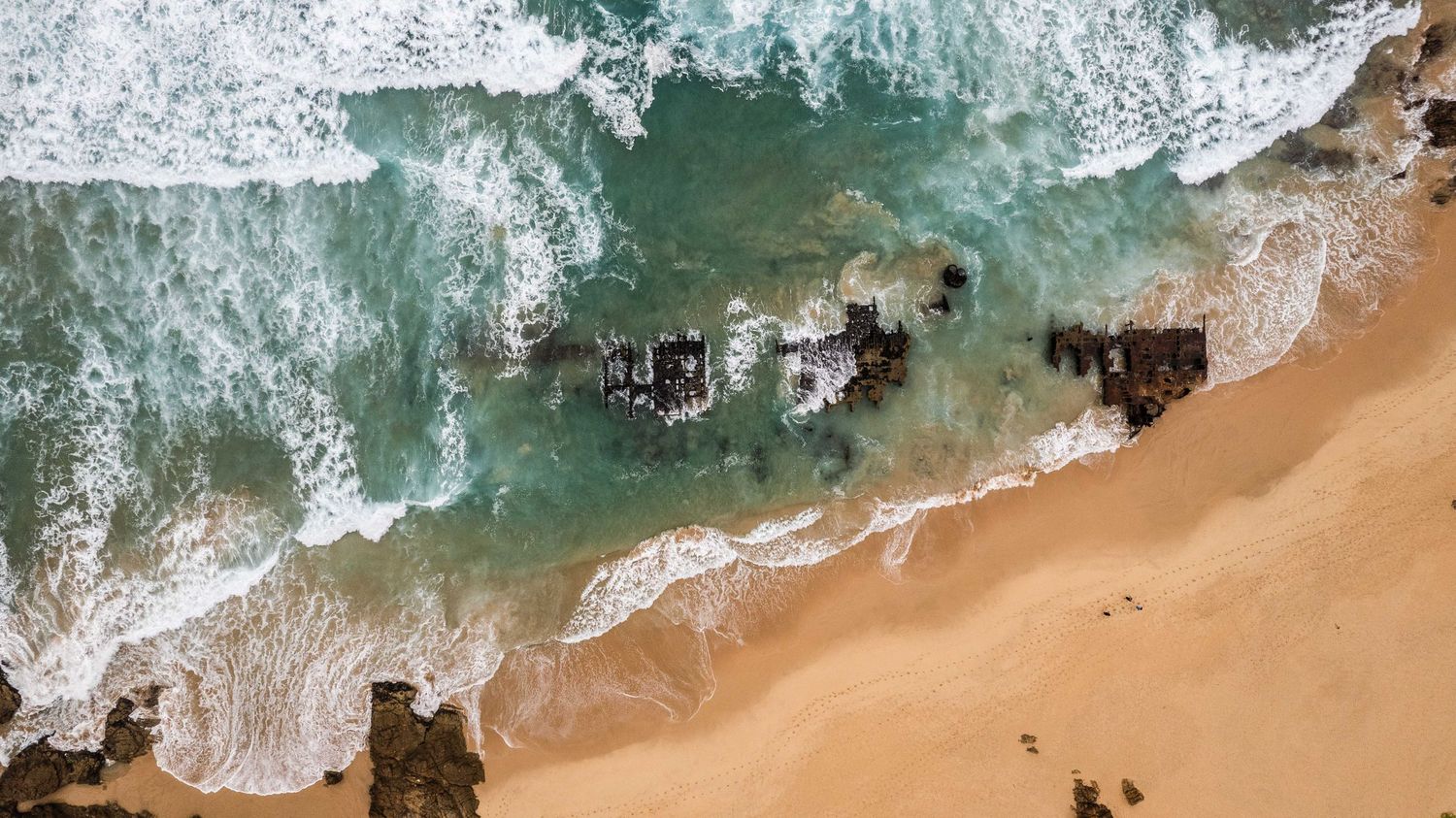 Aerial view of turquoise ocean waves crashing on a sandy beach with white foam patterns.