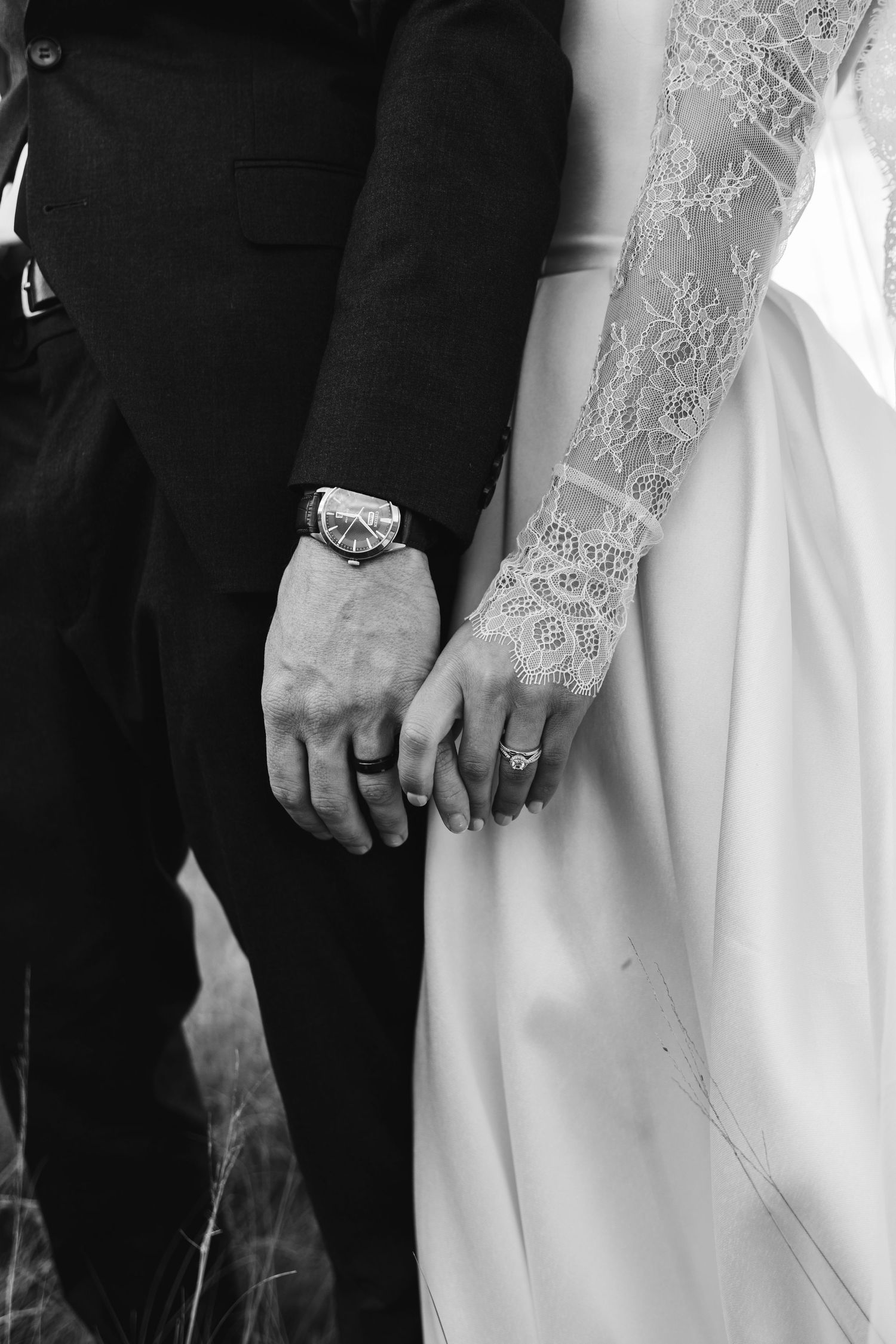 Black and white photo of bride and groom holding hands, showing lace wedding dress sleeve and groom watch.
