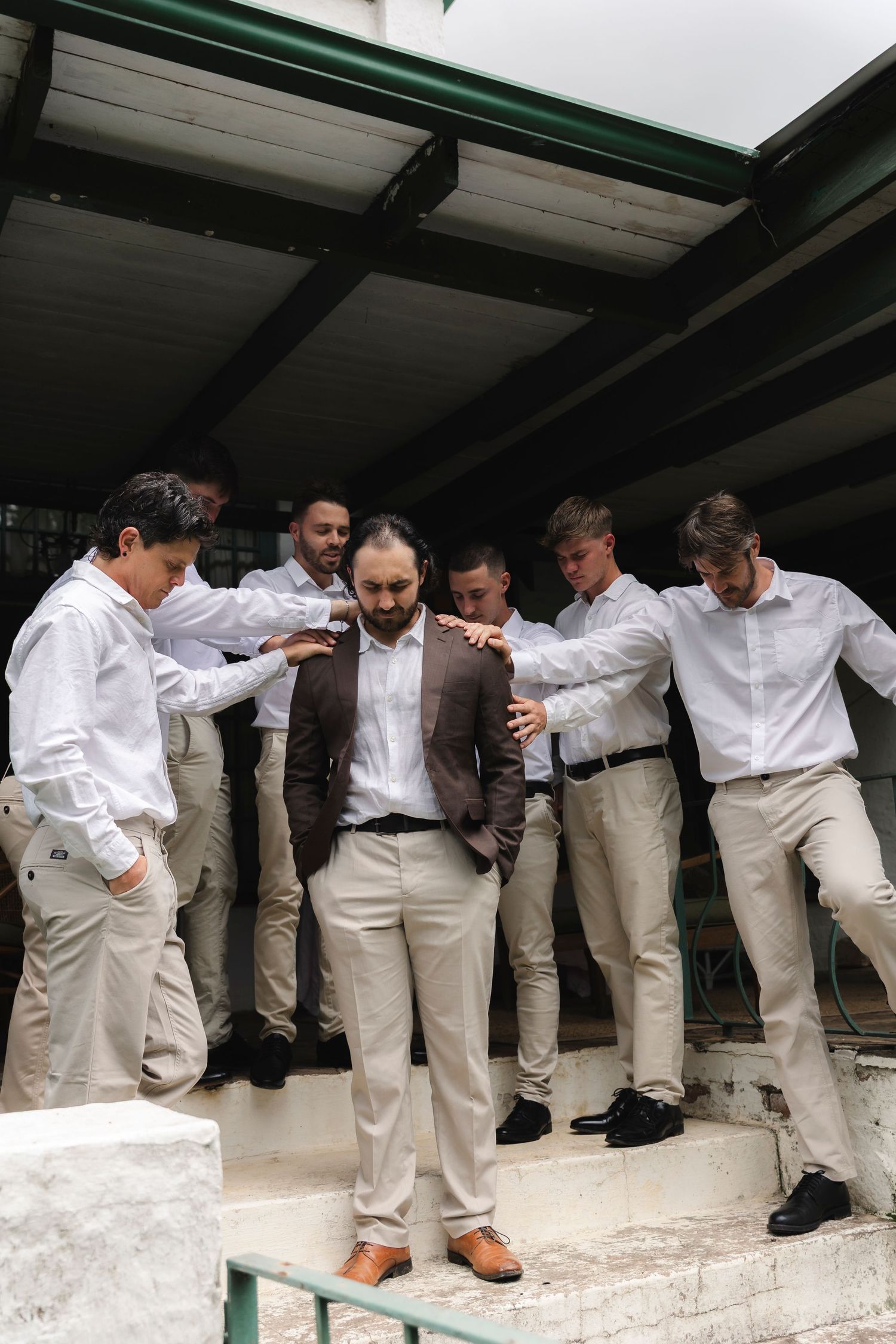 Groom in brown jacket surrounded by groomsmen in white shirts and khaki pants on steps during wedding day preparations.