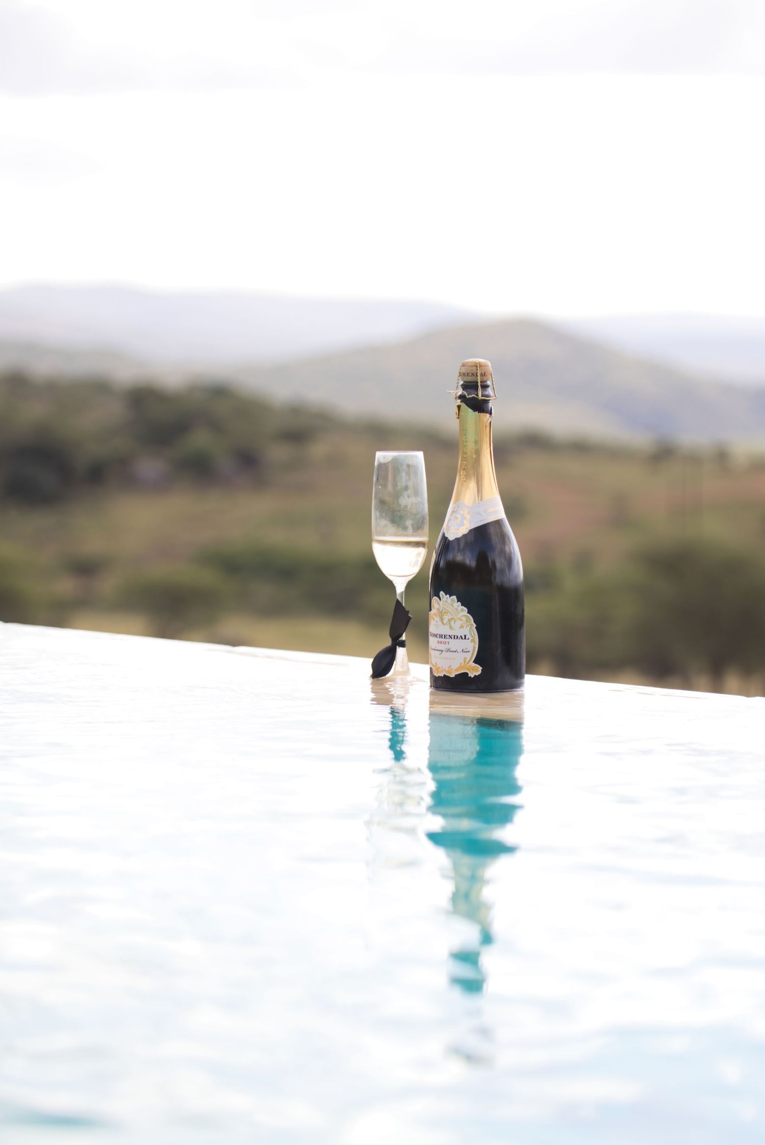 Champagne bottle and flute glass resting on infinity pool edge with scenic mountain landscape in background.