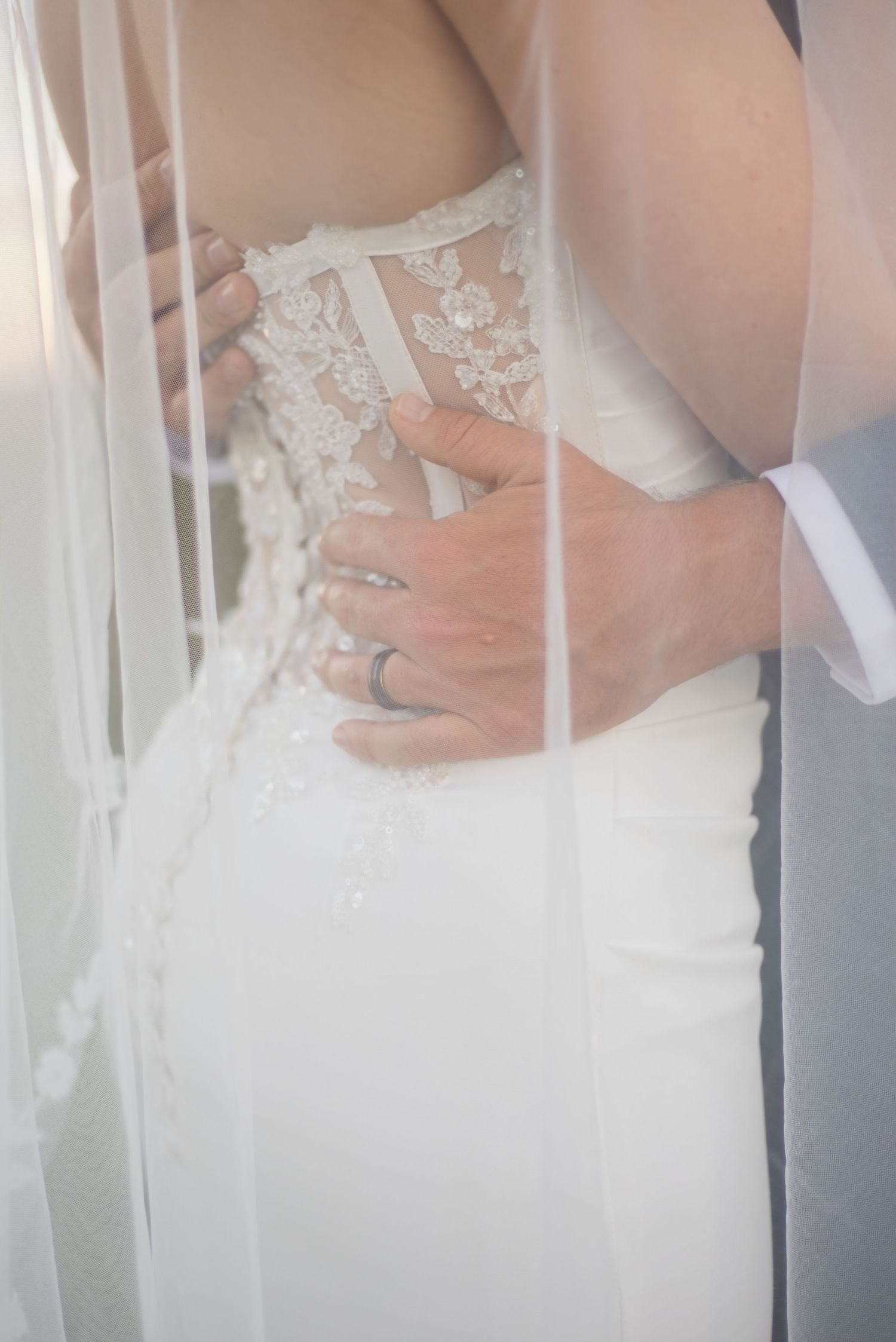 Groom embracing bride from behind, showing lace wedding dress back detail and wedding ring through sheer veil.