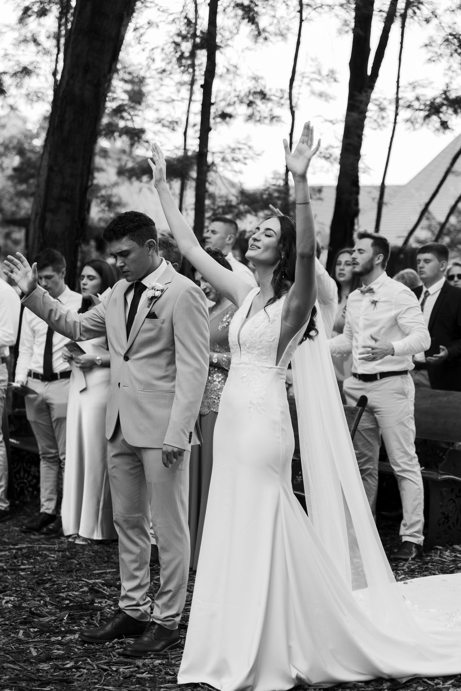 Black and white photo of a joyful bride raising her arms beside groom, surrounded by wedding guests in a forest setting.