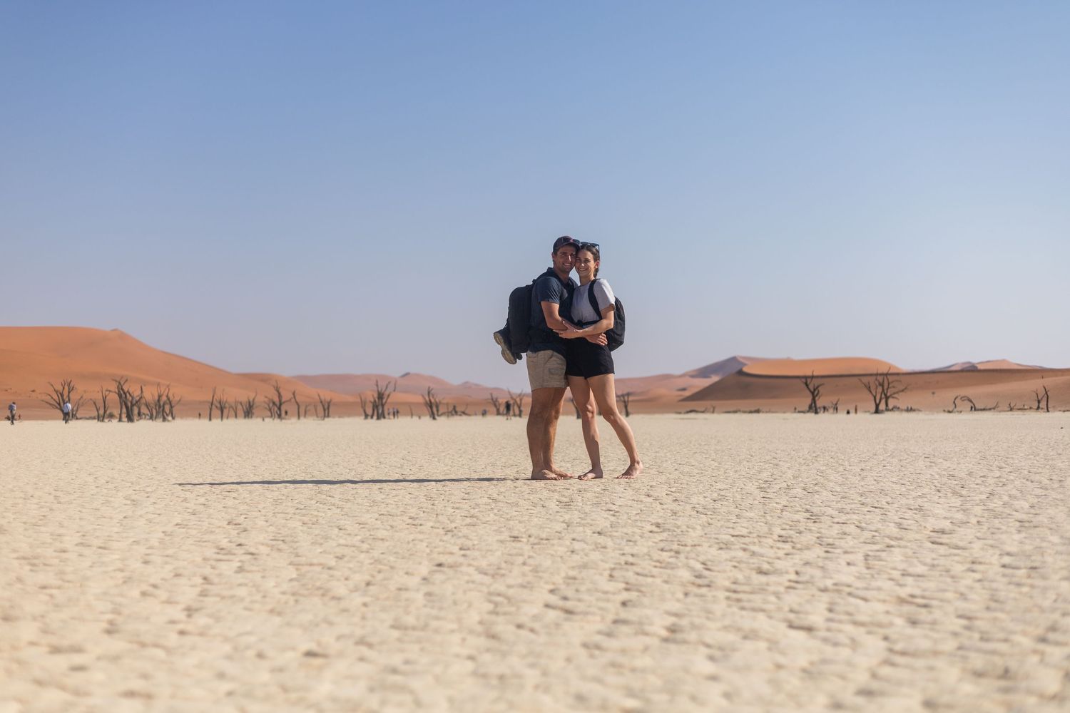 A couple shares an intimate moment in front of towering orange sand dunes in the desert.