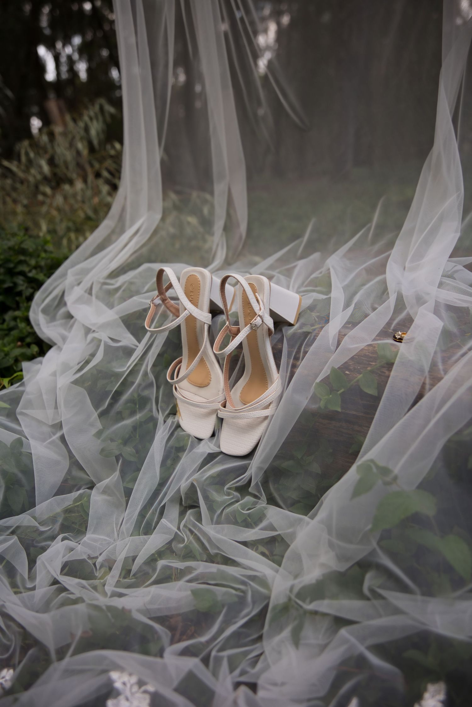 White bridal heels with ankle straps placed on flowing sheer tulle veil outdoors.