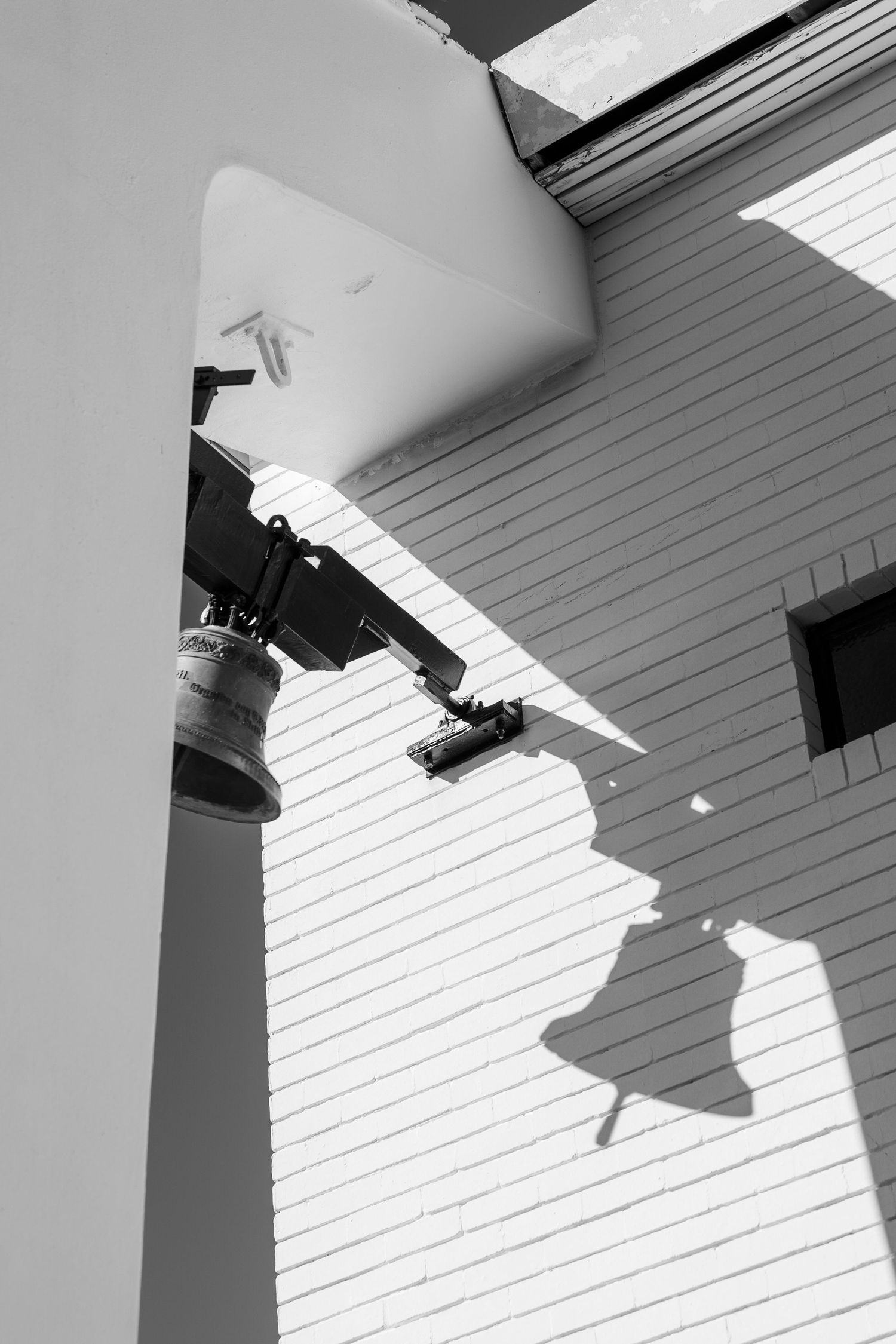 Black and white photo of a bell casting a shadow on a wooden building exterior with a security camera mounted above.