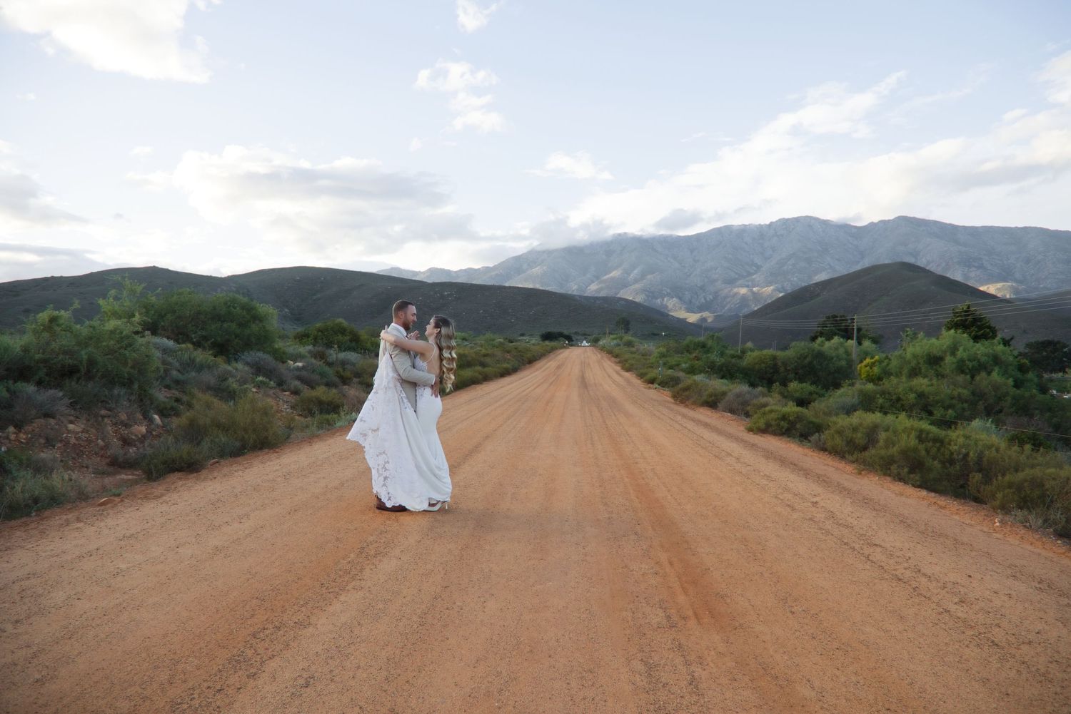 Two brides in white gowns embrace on a scenic dirt road surrounded by mountains and desert shrubs under a cloudy sky.