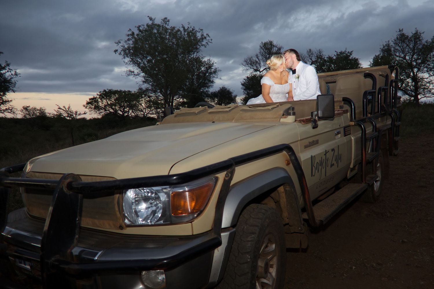 Bride and groom posing on a safari jeep at dusk with dramatic cloudy skies in the background.