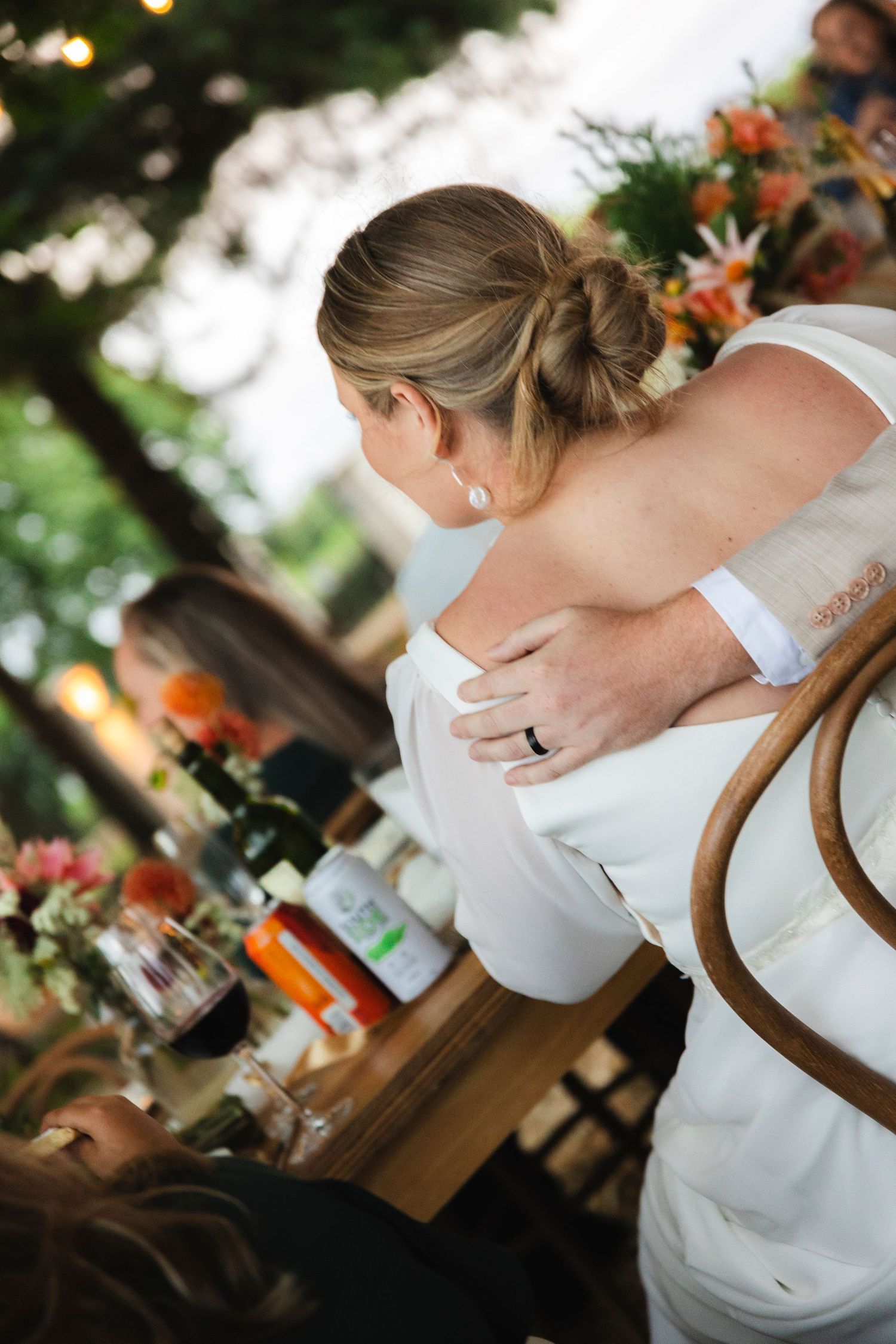 Bride in white gown seated at outdoor wedding reception table with floral arrangements and guests in background.